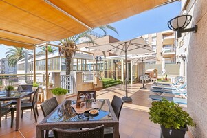 Un espacio de patio al aire libre con mesas y sillas, una mesa con superficie de vidrio, plantas en macetas y sombrillas que dan sombra.