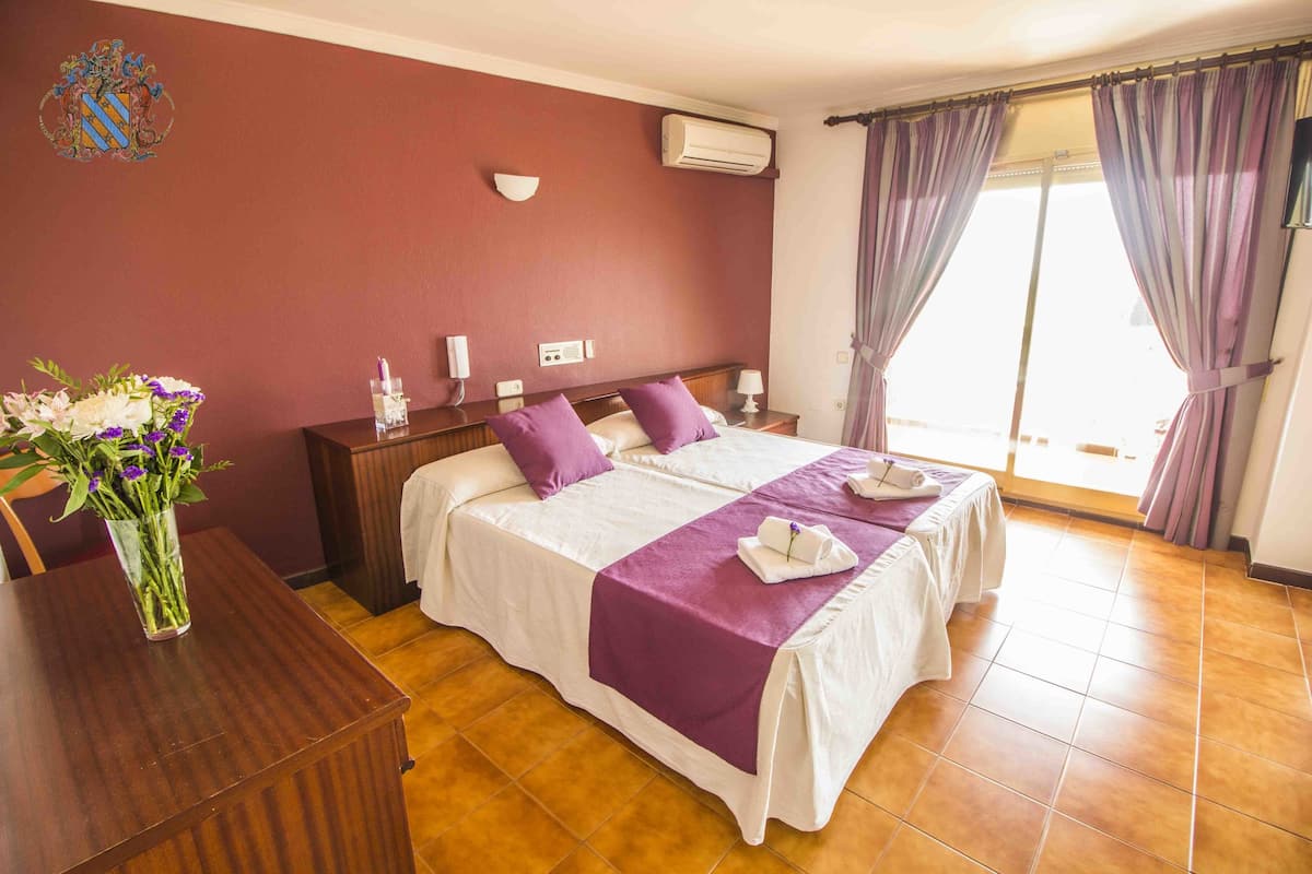A hotel room with two beds, a wooden desk, a vase of flowers, and a window with curtains.
