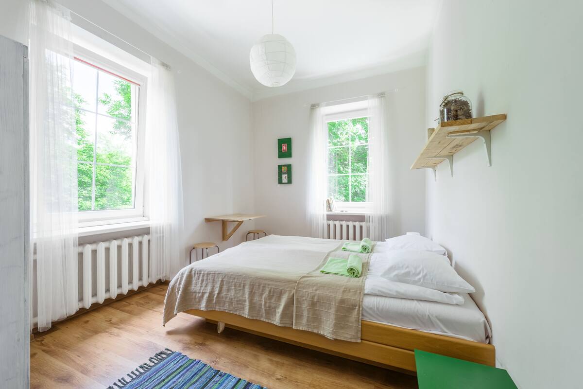 Un dormitorio con cama de madera, dos ventanas con cortinas, un radiador, una mesita y una lámpara colgante.