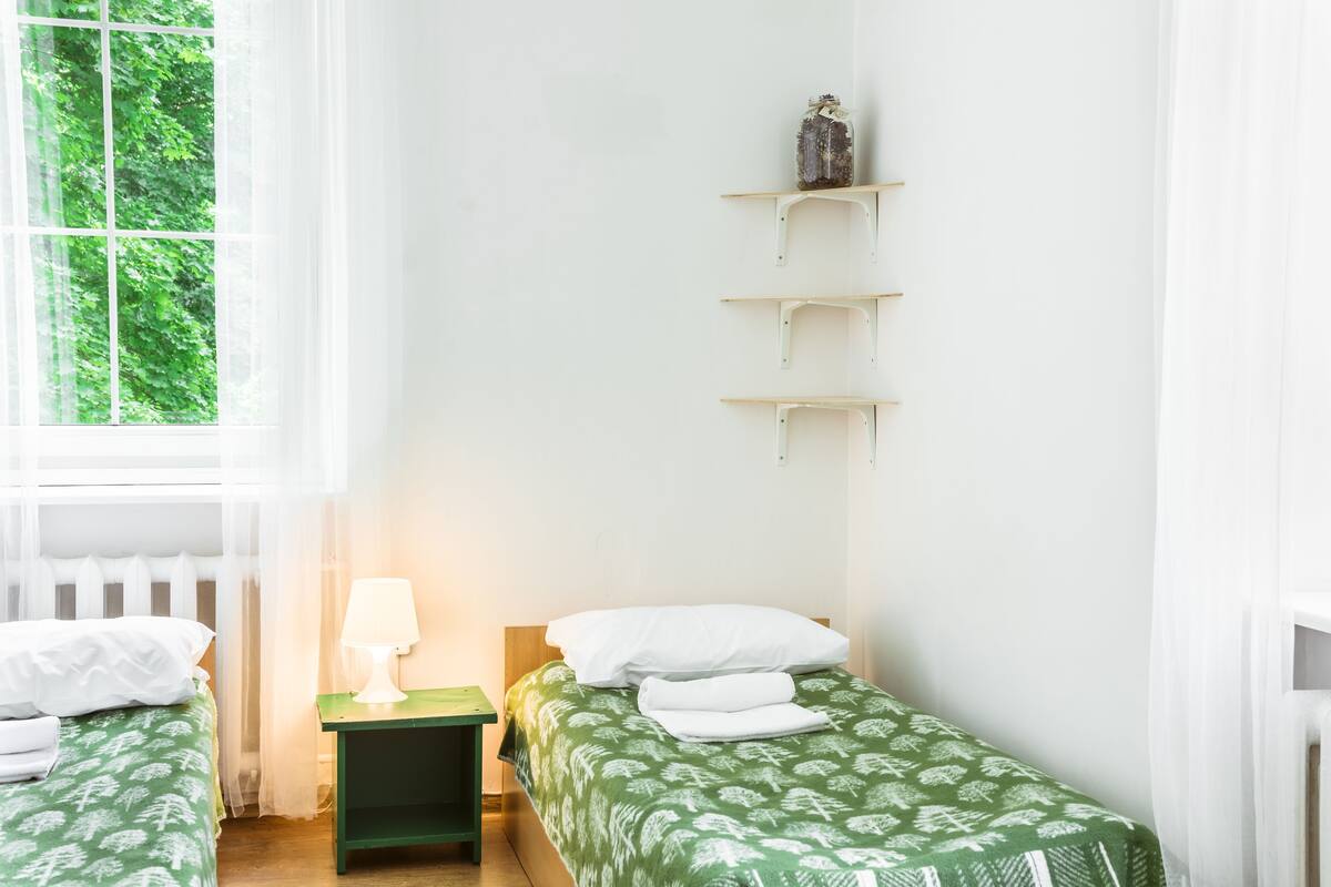 Un dormitorio con dos camas individuales, una mesita de noche con lámpara y una ventana con vistas a la vegetación.