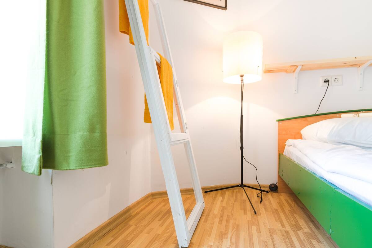Una habitación con suelo de madera, cama con ropa de cama verde y blanca, una lámpara de pie y un cuadro en la pared.