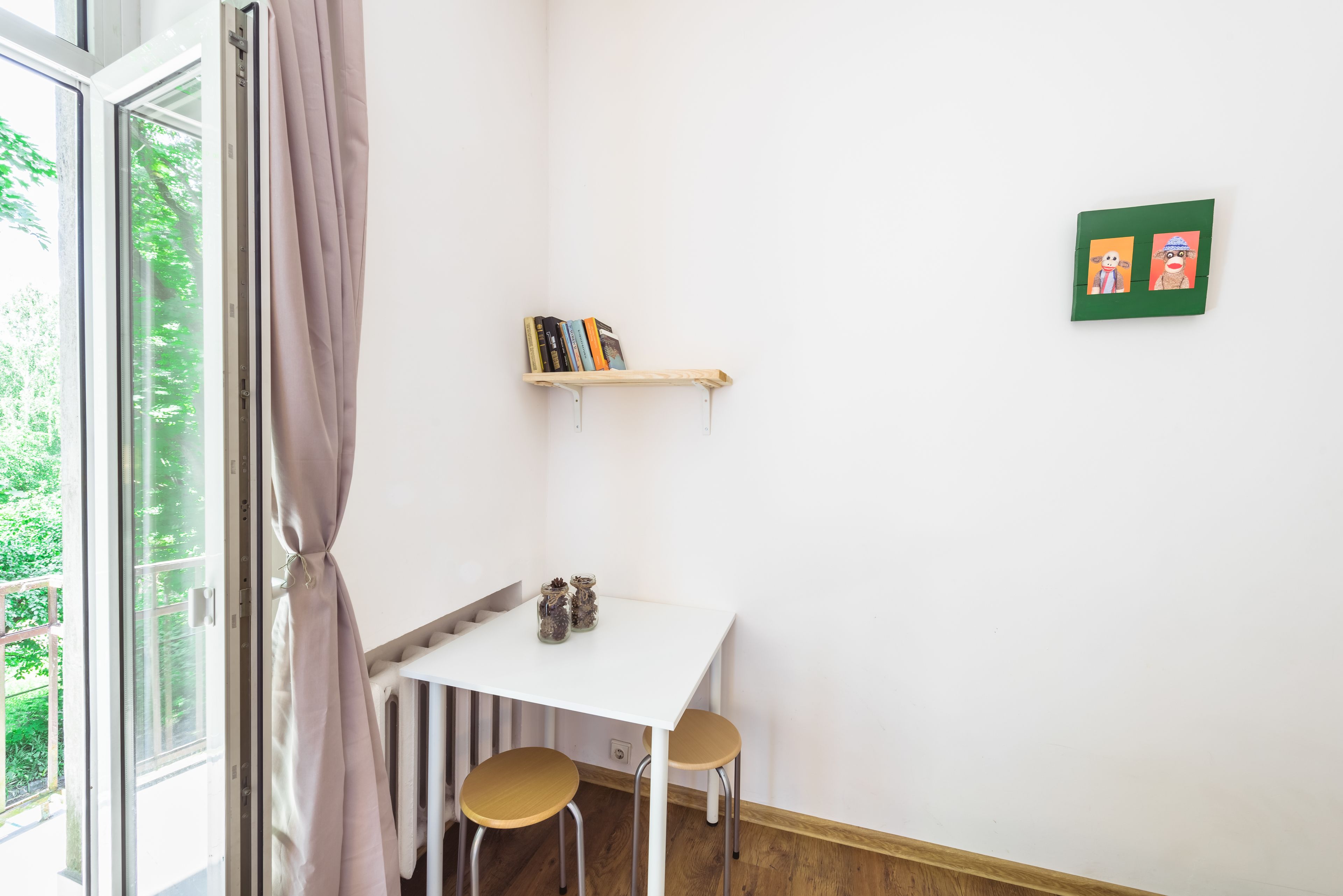 A small room with a white table, two stools, a bookshelf, and a window with curtains.