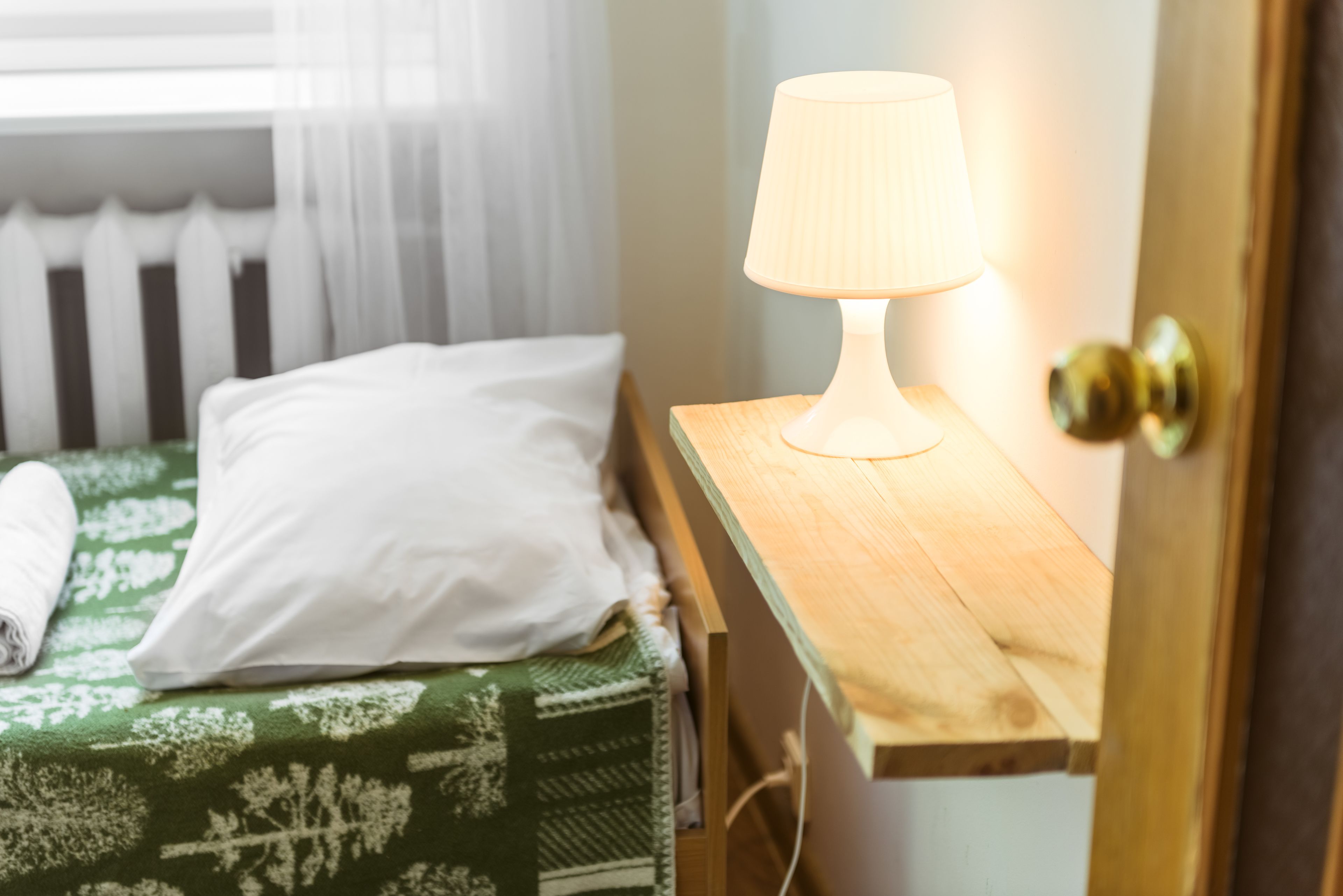 A neatly made bed with a green patterned bedspread and white pillows, a bedside table with a lamp, and a wooden door with a brass doorknob.