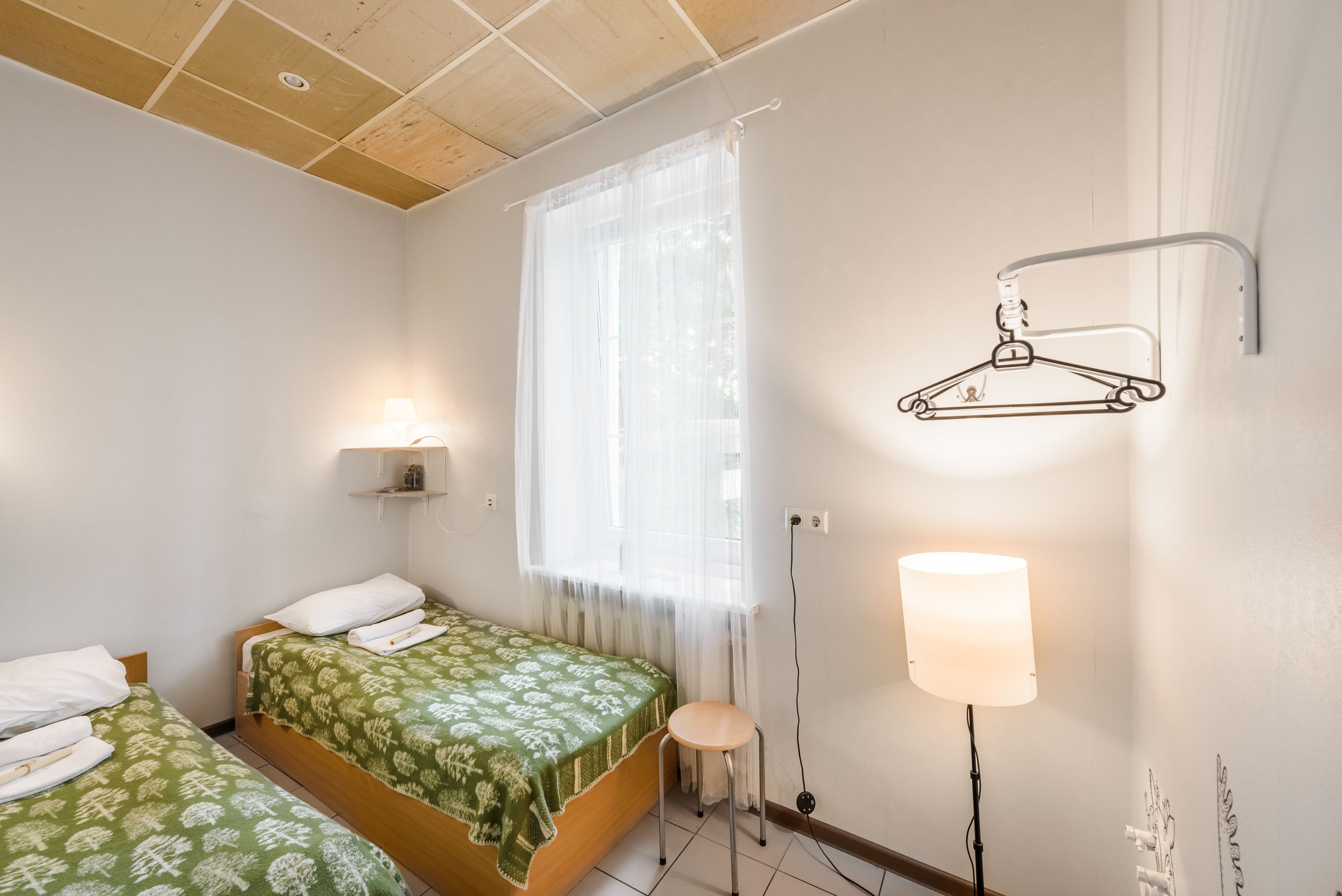 Un dormitorio con dos camas, una ventana con cortinas translúcidas, un perchero en la pared, una mesita de noche con lámpara y un pequeño estante con un objeto decorativo.