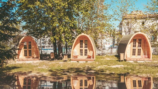 Three wooden arched cabins by a reflective water body, with a large tree and a building in the background. - Downtown Forest Hostel & Camping (Vilnius)