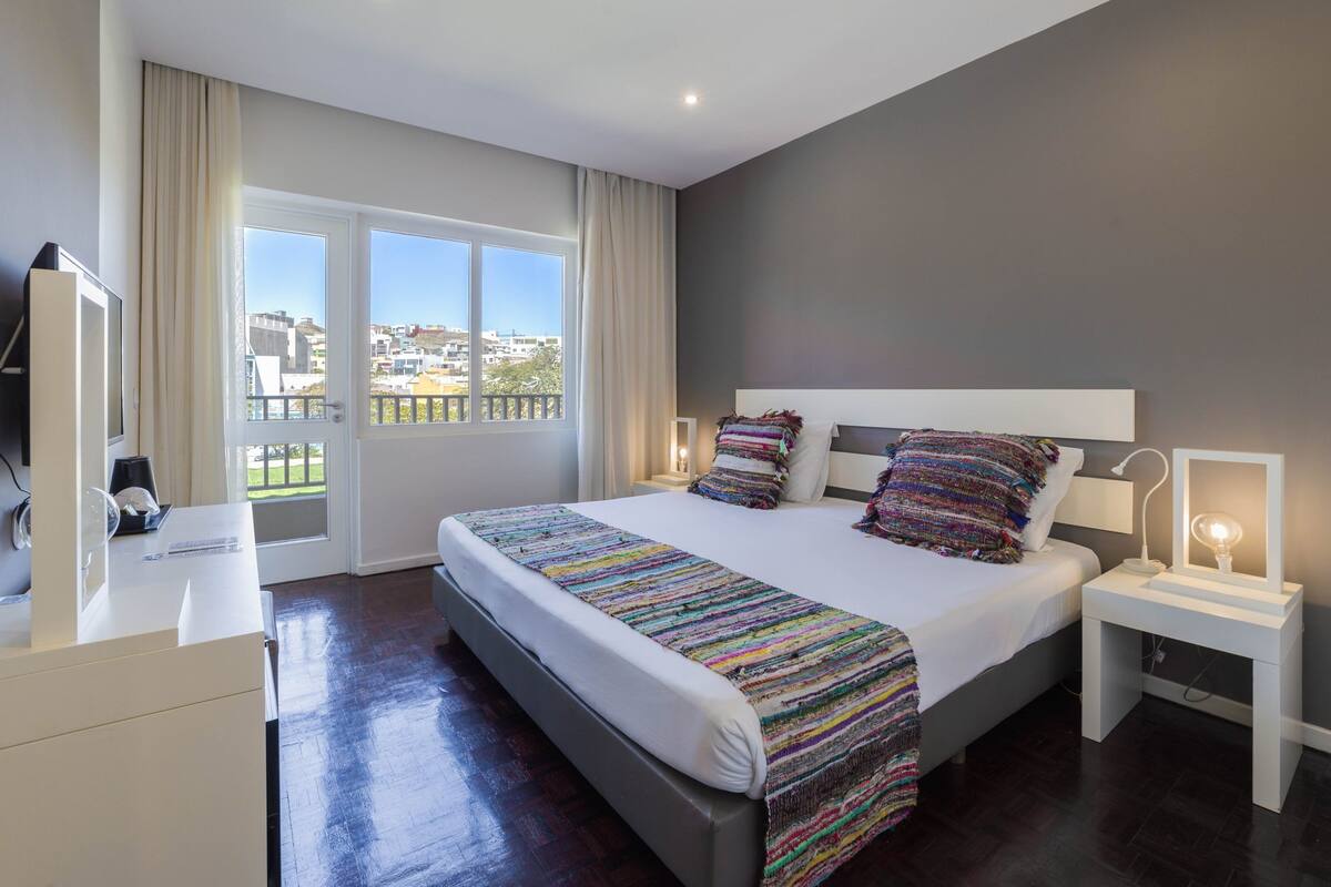 Une chambre d’hôtel moderne dotée d’un grand lit, de tables de chevet, d’une télévision et d’un balcon offrant une vue sur la ville.