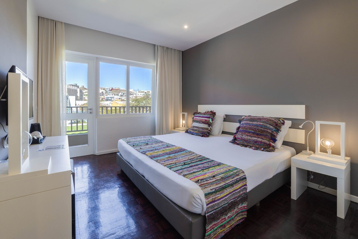 Ein modernes Hotelzimmer mit einem großen Bett, Nachttischen, einem Fernseher und einem Balkon mit Blick auf die Stadt.
