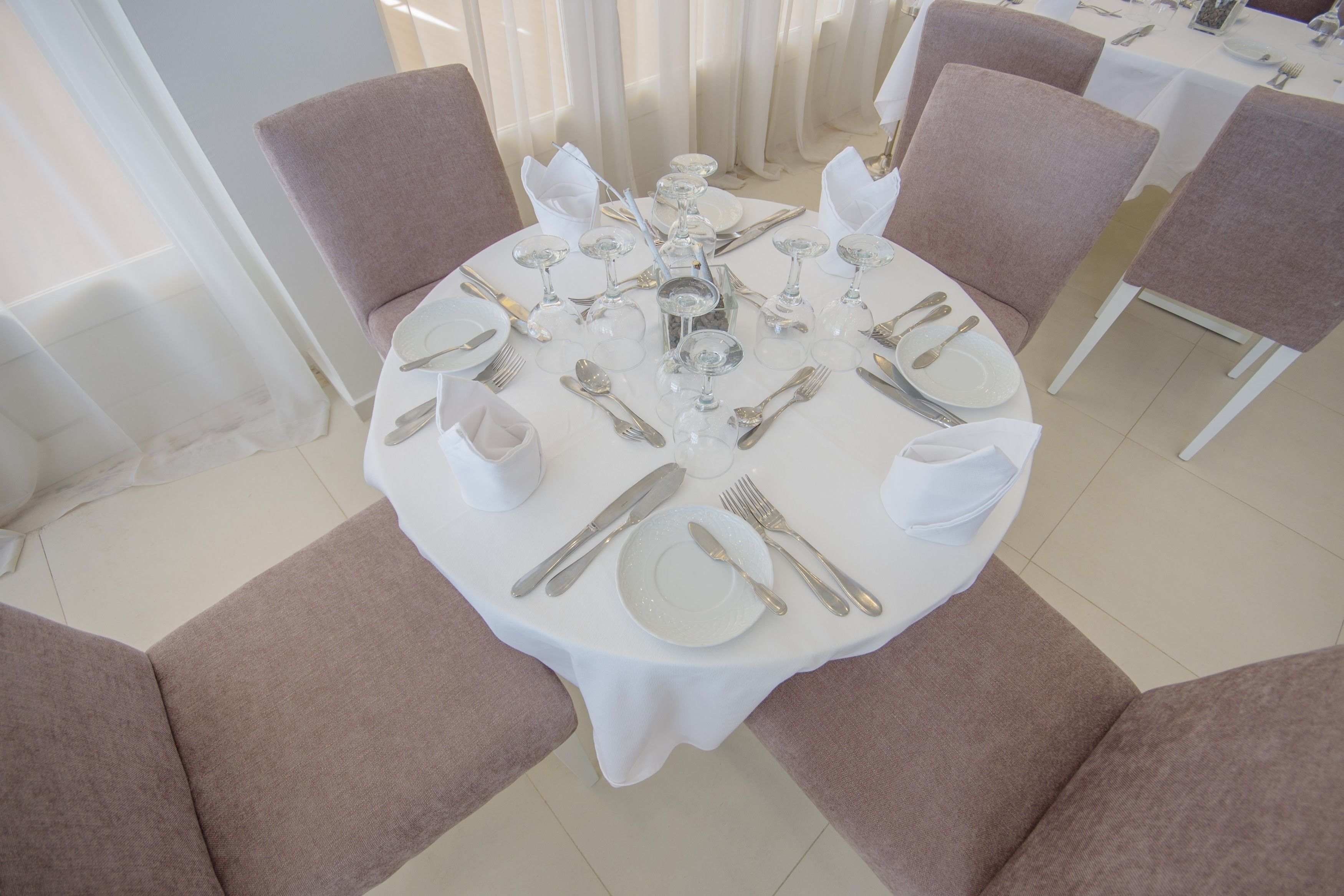 Mesa redonda para comedor con mantel blanco, cubiertos y cristalería, rodeada de sillas con cojines.