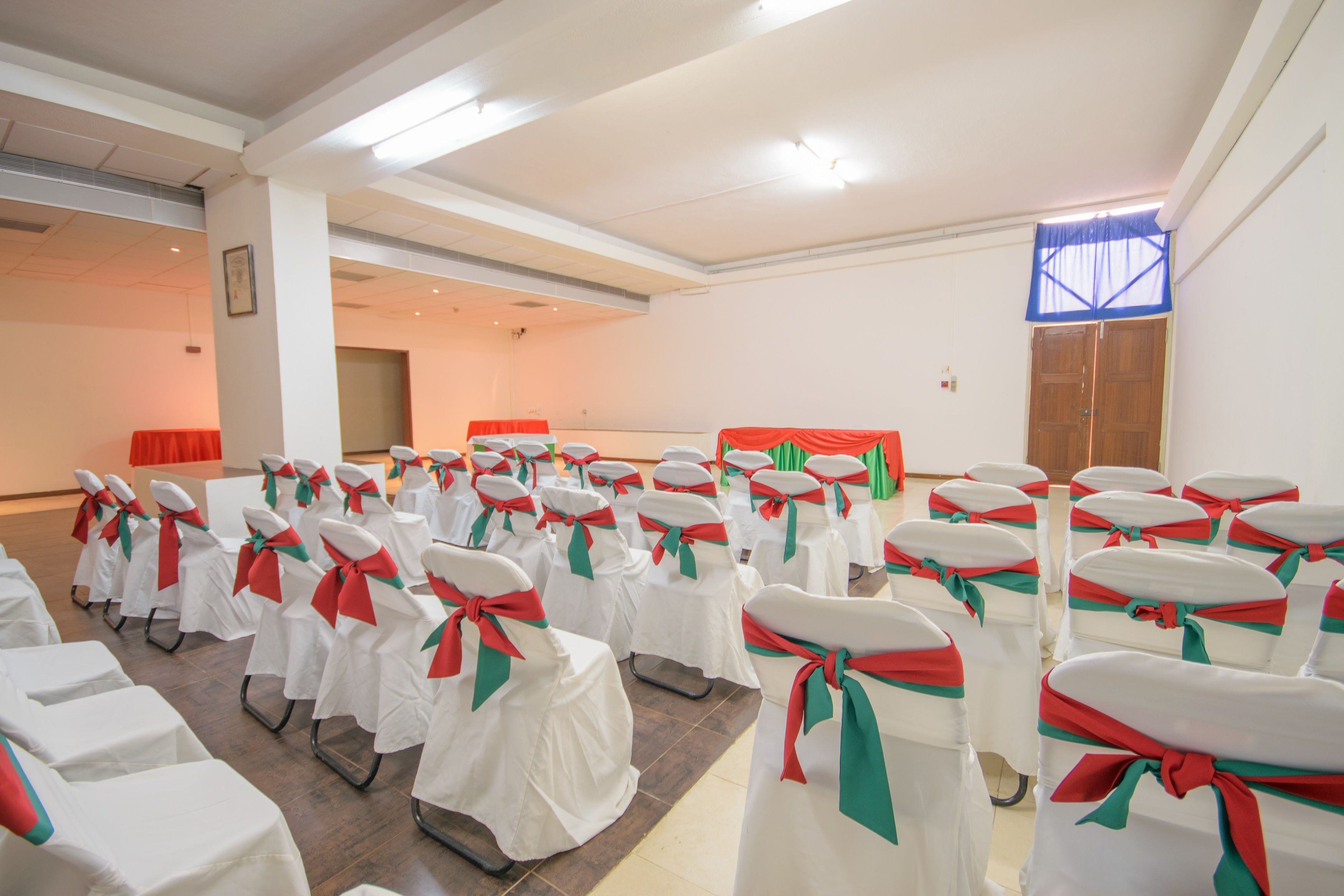 A conference room with rows of white chairs with red and green sashes, a stage with a red backdrop, and a large window with blue curtains.