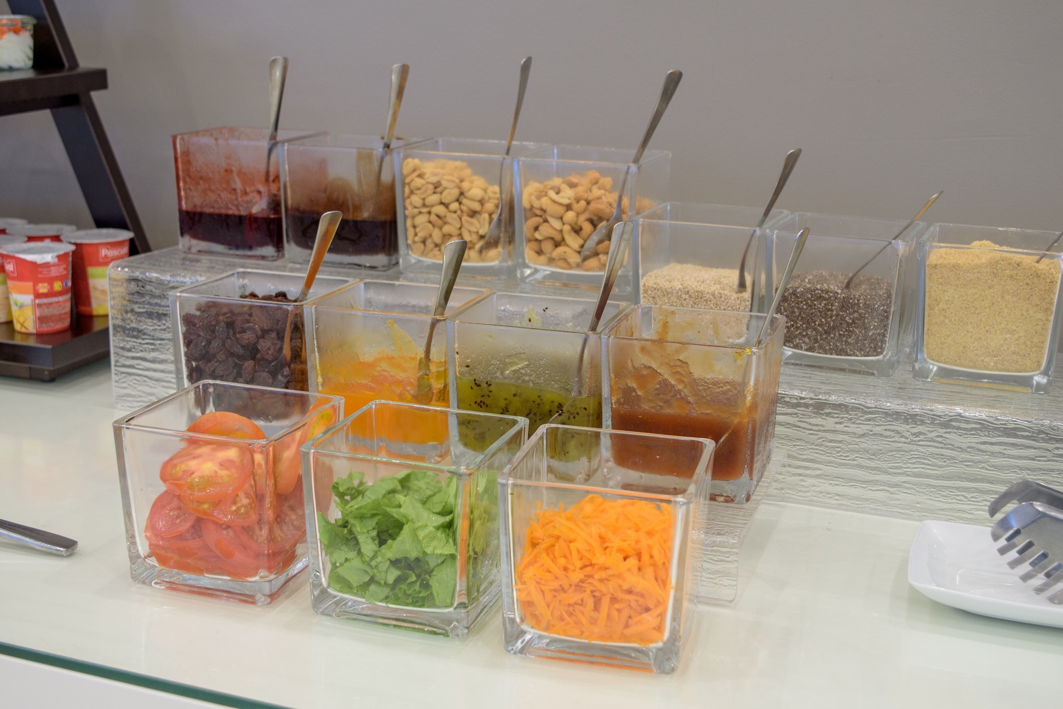 Un bufé con diversos alimentos en recipientes transparentes, cada uno con una cuchara.