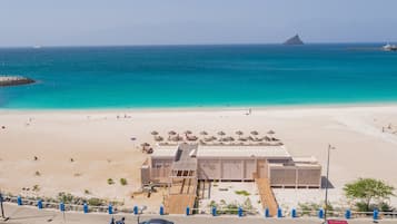 Un bâtiment situé au bord de la plage, doté d’un toit plat, d’une promenade pavée menant à la plage et offrant une vue dégagée sur l’océan et une île lointaine.