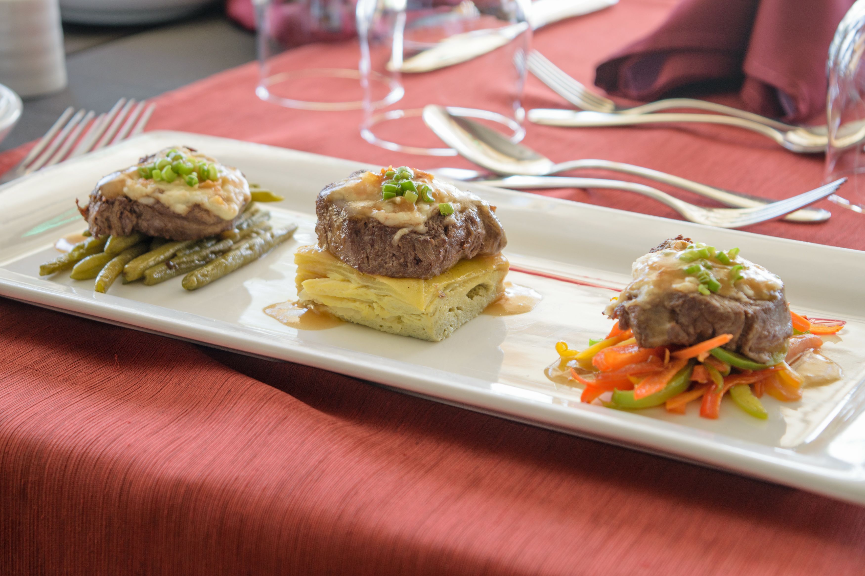 Une assiette rectangulaire blanche présente trois plats de viande, chacun nappé d’une sauce crémeuse et garni d’oignons verts, le tout servi sur une nappe rouge.