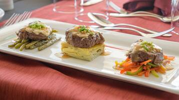 Une assiette rectangulaire blanche présente trois plats de viande, chacun nappé d’une sauce crémeuse et garni d’oignons verts, le tout servi sur une nappe rouge.