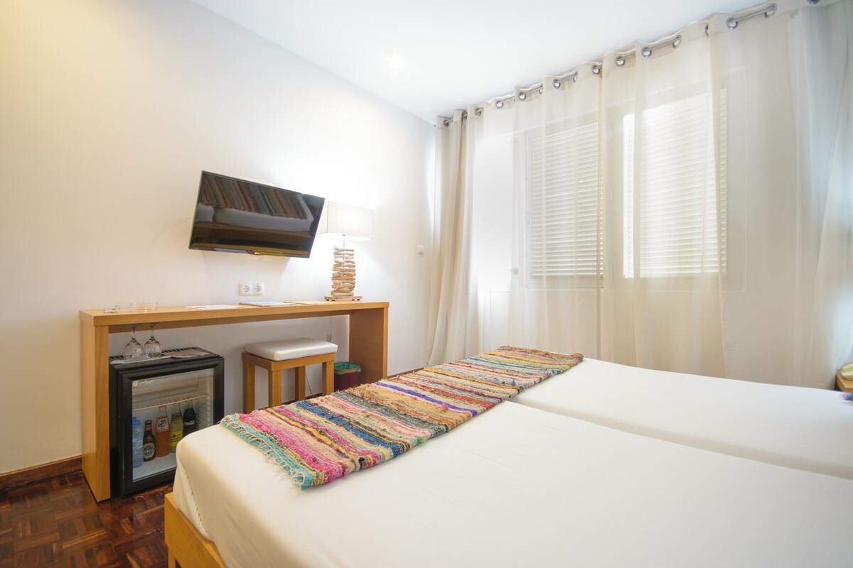 Ein Hotelzimmer mit Bett, Schreibtisch mit Fernseher, Minikühlschrank und einem Fenster mit Vorhängen.