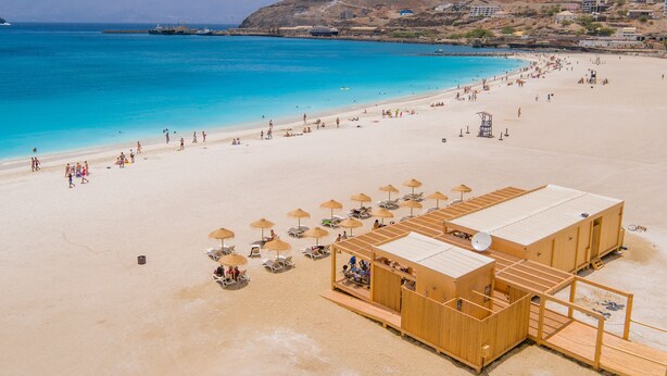 Een strand met een houten constructie, parasols en stoelen, een heldere blauwe zee en bergen op de achtergrond.