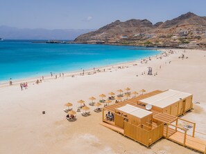 Una playa con una estructura de madera, sombrillas y sillas, un mar azul cristalino y montañas al fondo.