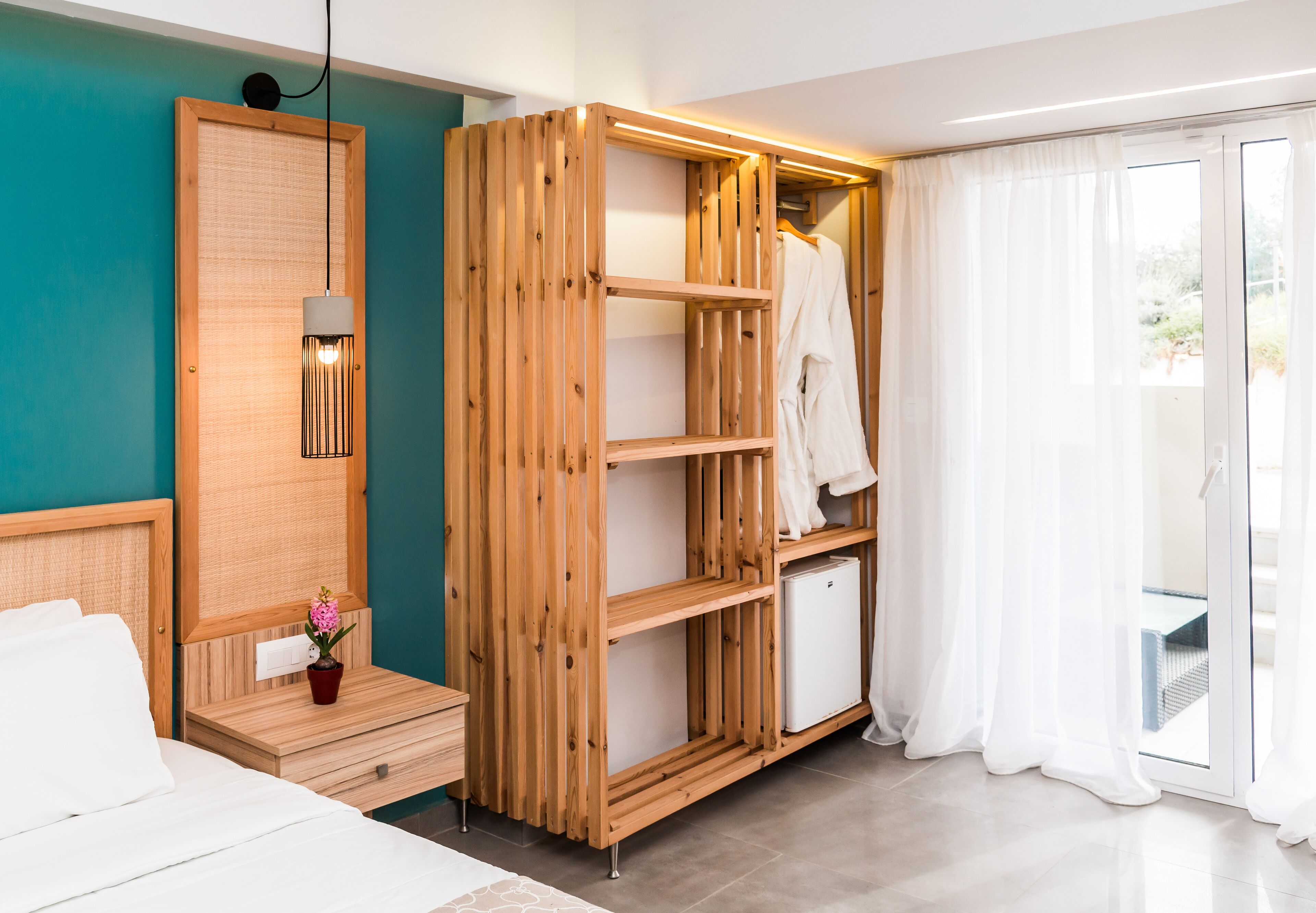 Ein modernes Hotelzimmer mit einem Holzschrank, einem kleinen Tisch mit einem Topfpflanzen und einem Bett mit weißer Bettwäsche sowie einer Schiebetür zum Außenbereich.