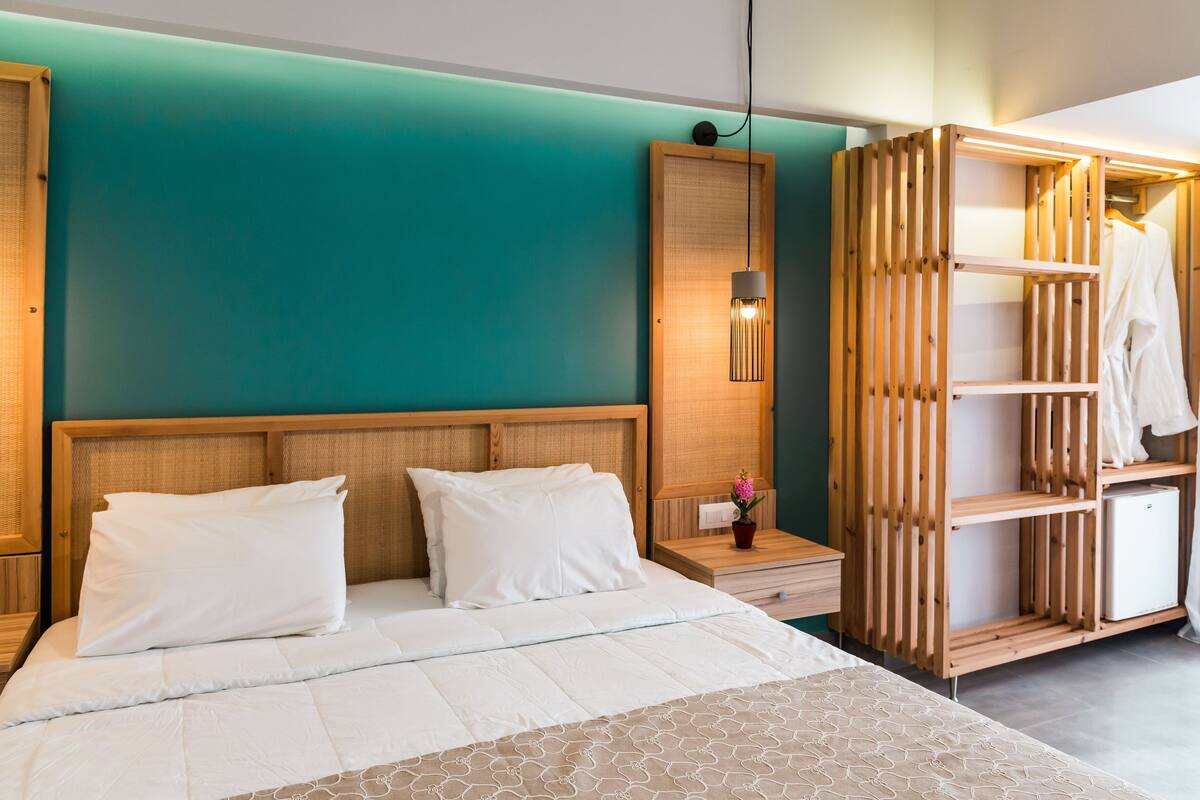 A modern hotel room with a teal accent wall, a wooden headboard, a bed with white linens, a nightstand with a lamp, and a built-in wardrobe with a hanging robe.