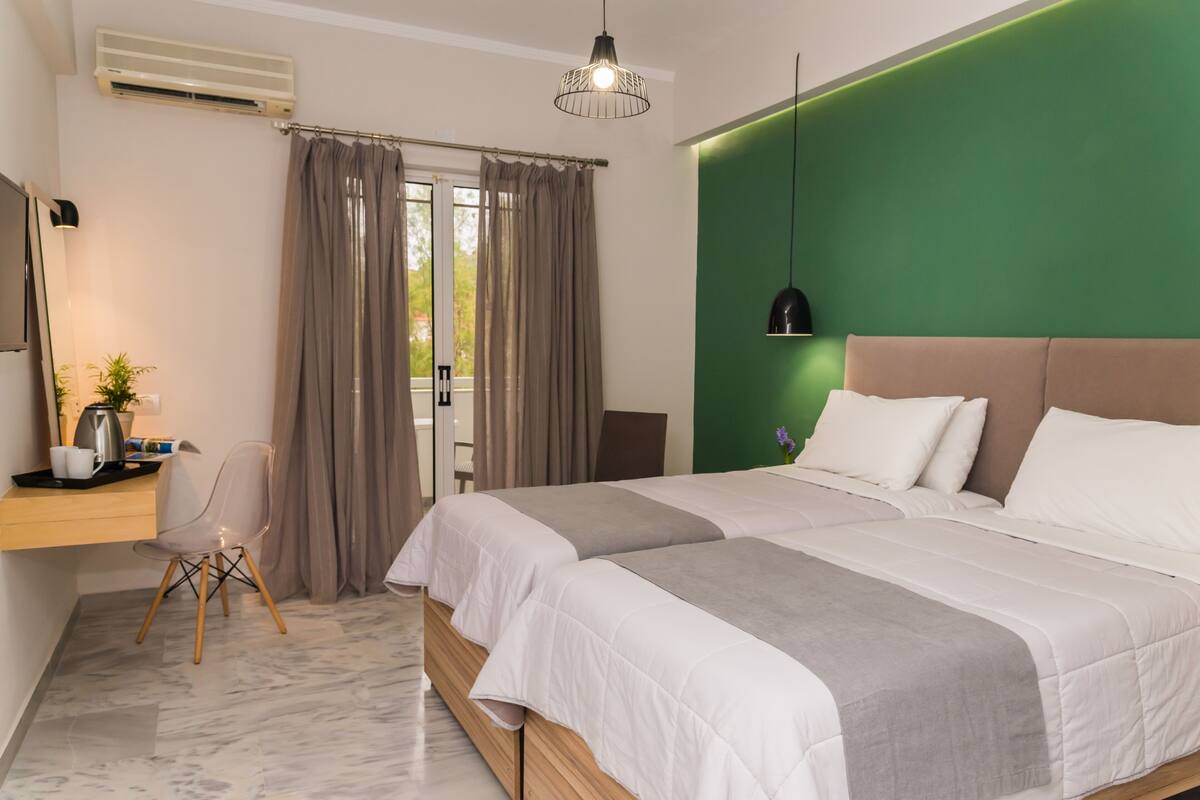A hotel room with two beds, a desk with a chair, a green accent wall, and a window with curtains.