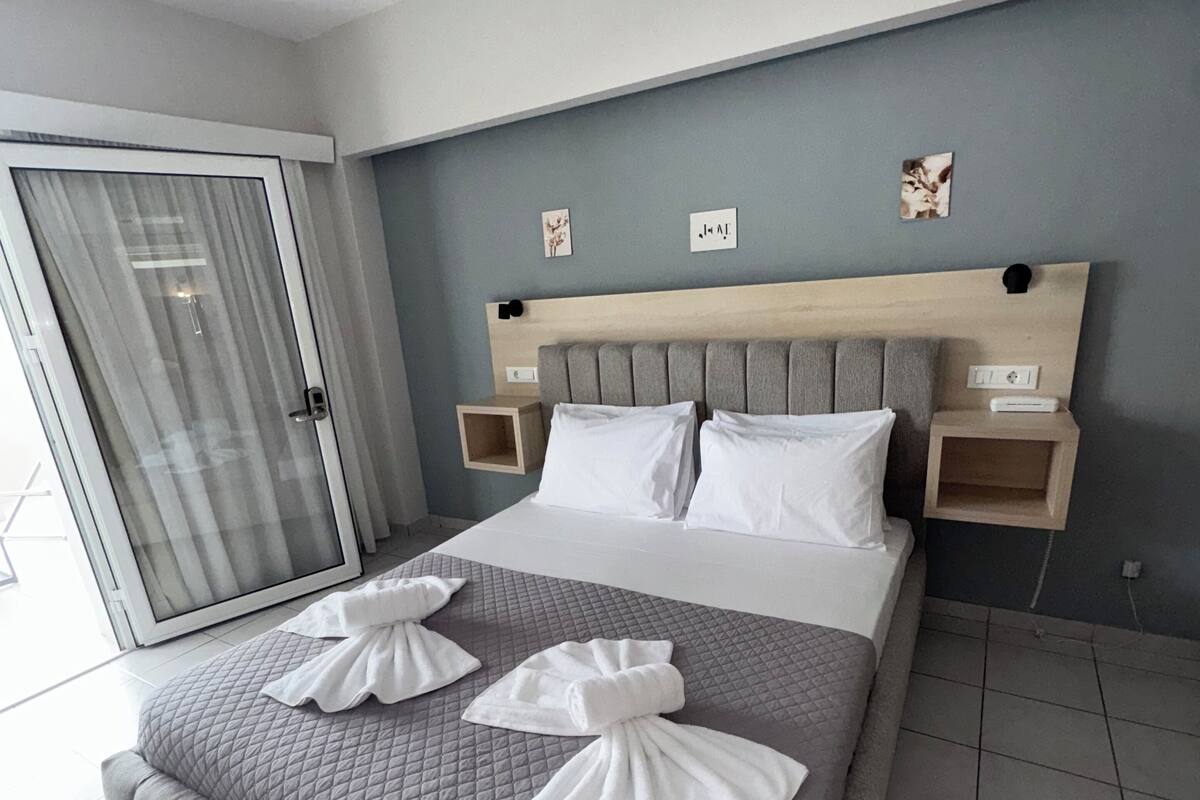 A modern hotel room with a large bed, grey quilted bedding, two bedside tables with lamps, and a sliding glass door leading to a balcony.