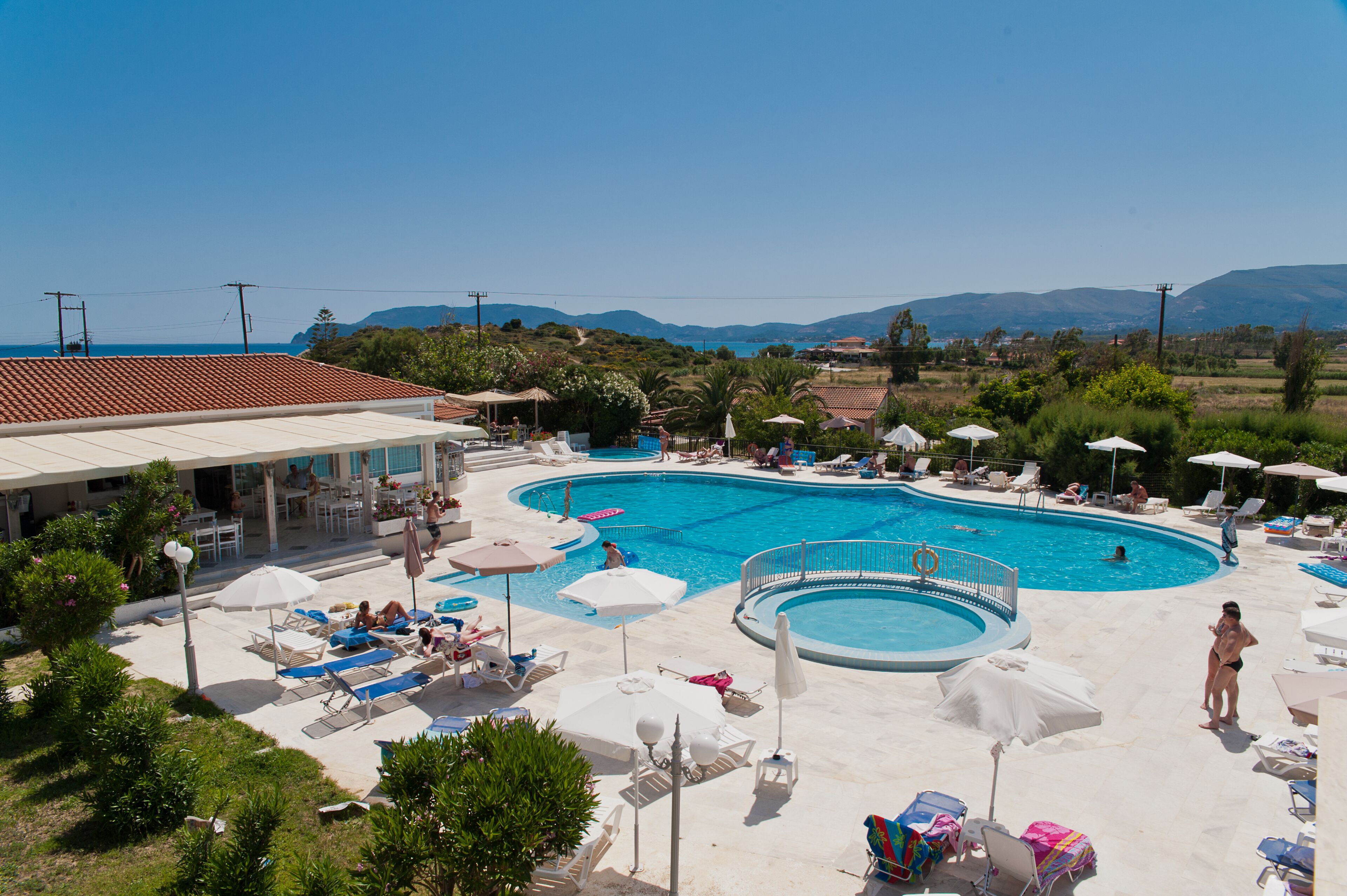 A large outdoor swimming pool with multiple sun loungers and umbrellas, surrounded by greenery and buildings.