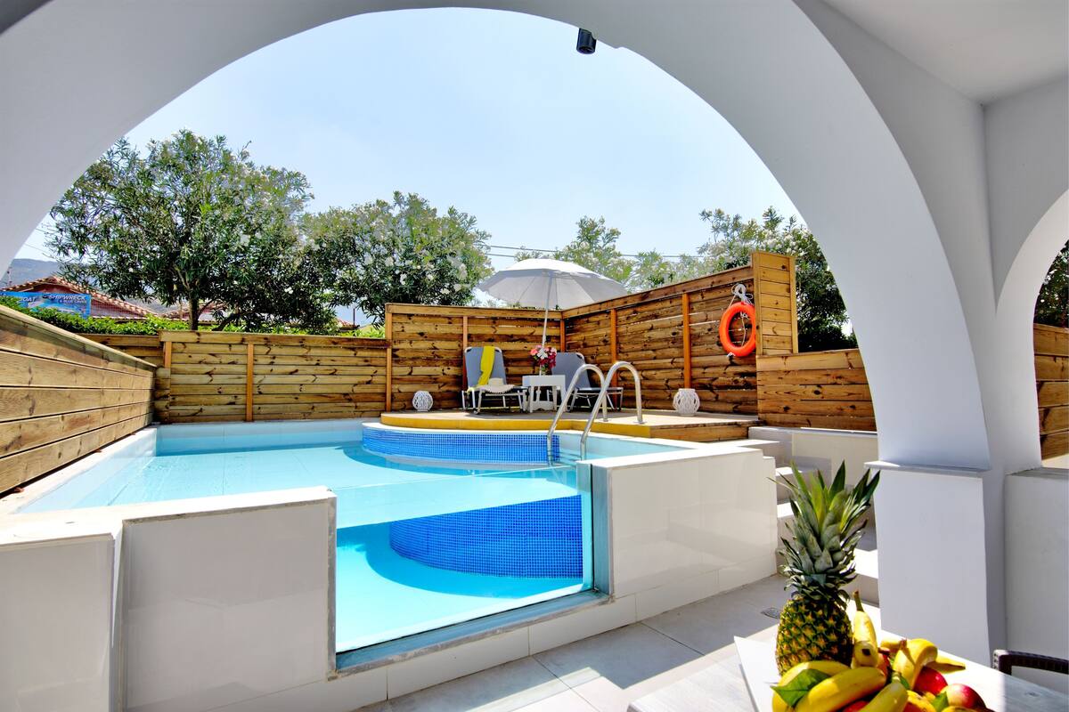 A swimming pool with a wooden deck, a white umbrella, and a fruit bowl.