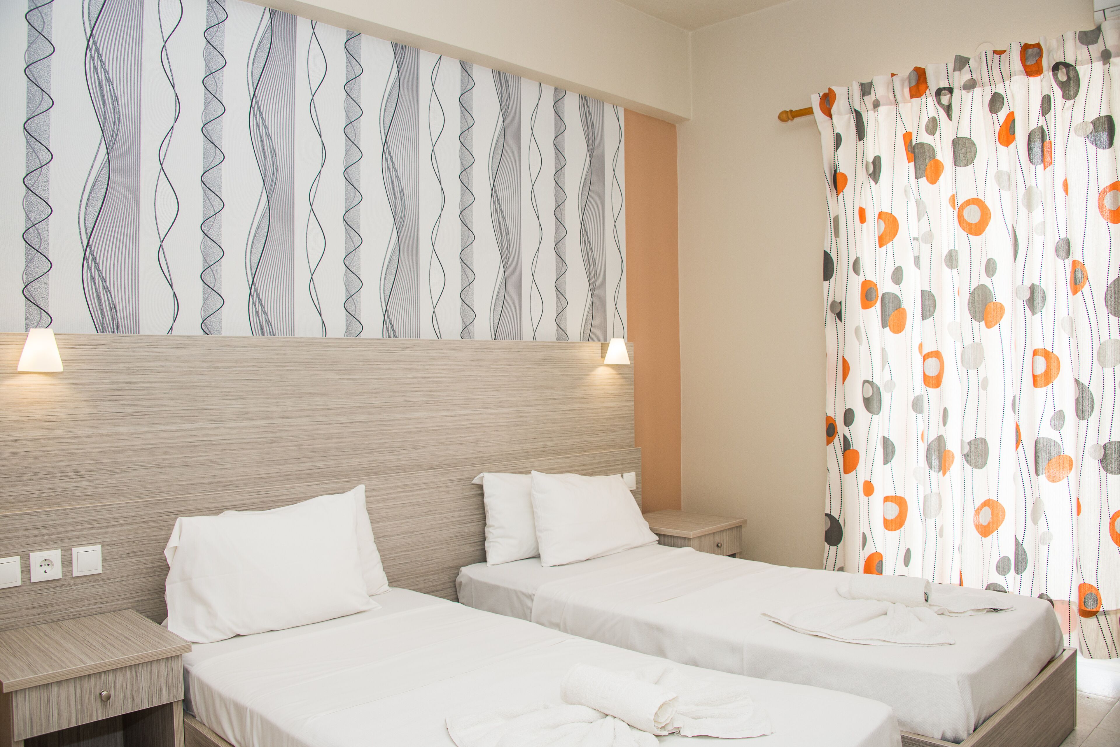 A hotel room with two single beds, a wooden headboard, a patterned wall, and a curtain with a geometric design.