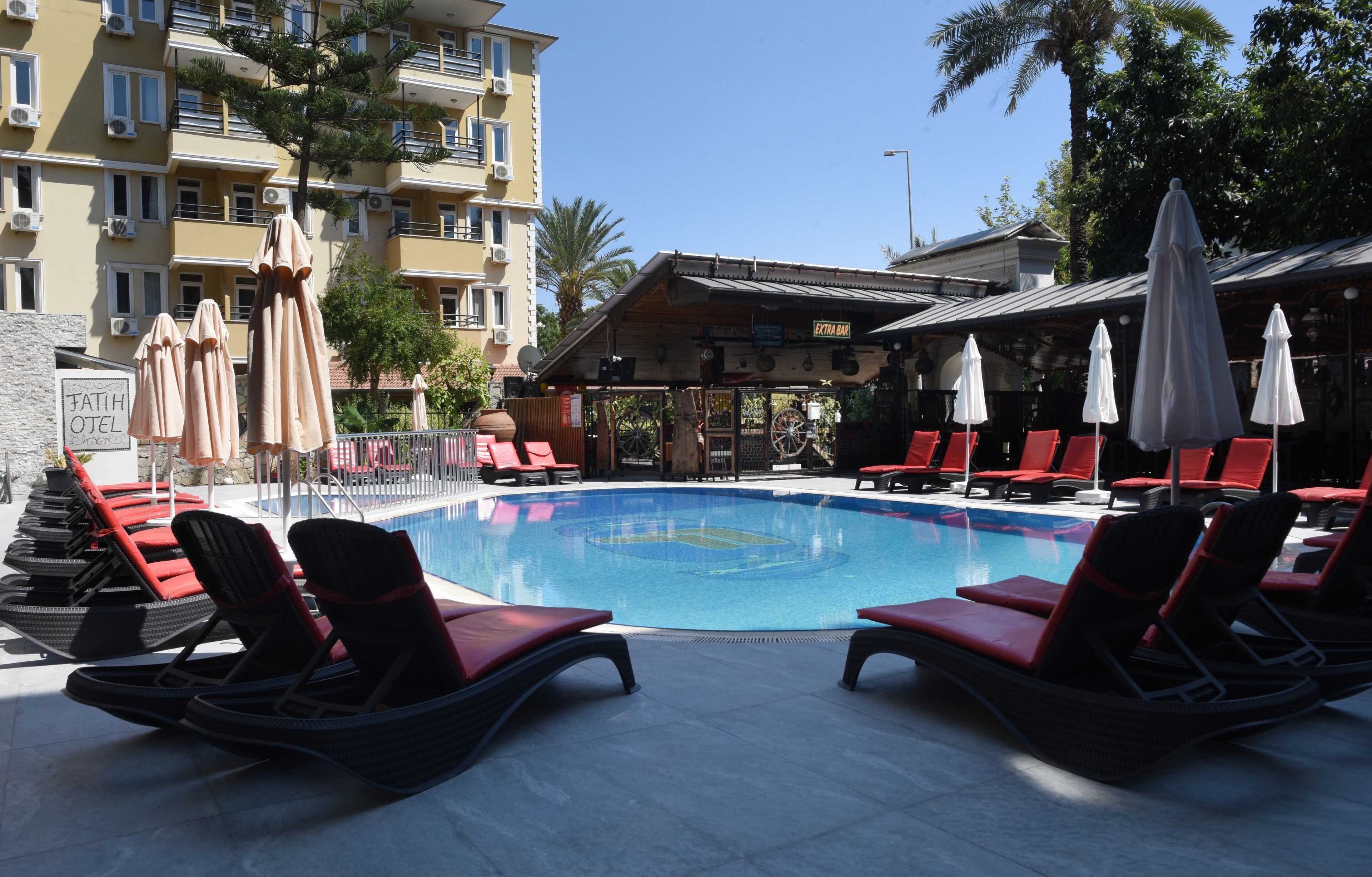Ein Swimmingpool mit roten Liegestühlen und Sonnenschirmen, umgeben von einem Gebäude mit Balkonen und Palmen.