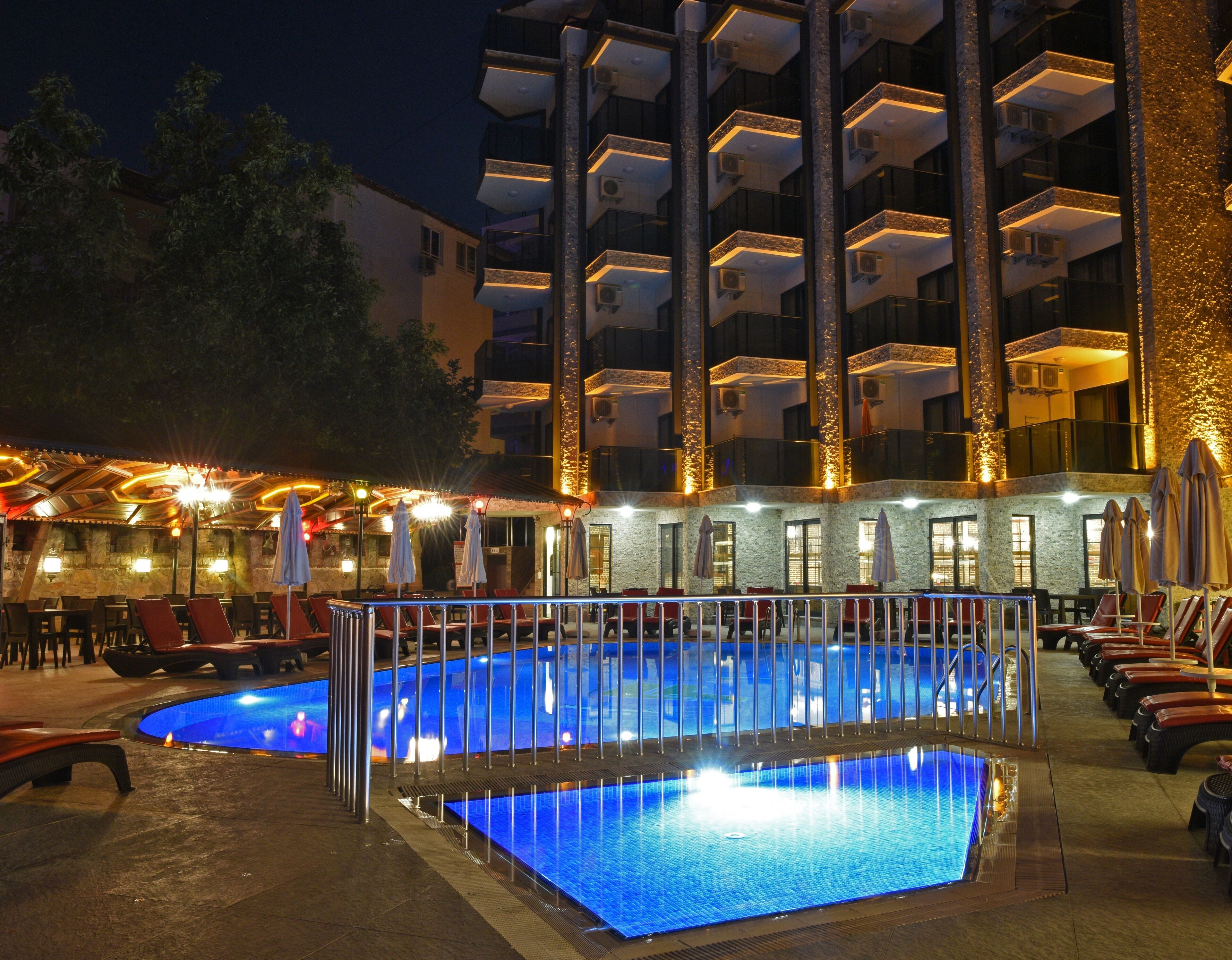 Una piscina con un deck circundante, sillas de descanso y un edificio con varios balcones al fondo.