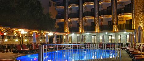 Una piscina con un deck circundante, sillas de descanso y un edificio con varios balcones al fondo.