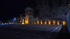 Um edifício de pedra histórico com uma torre, iluminado por luzes quentes, cercado por terreno coberto de neve e árvores despidas.