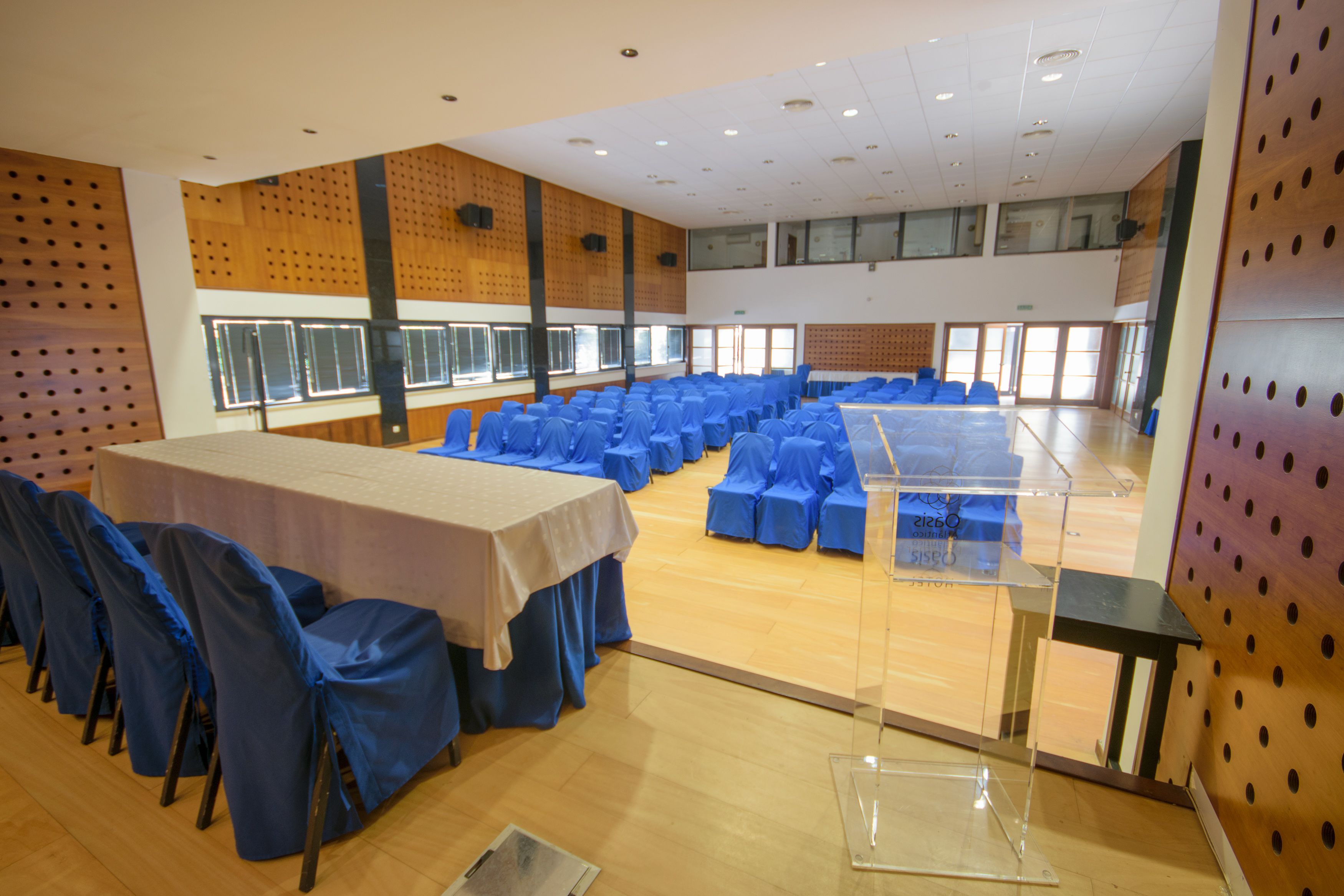 A large hall with rows of blue-covered chairs and a long table, wooden floors, and a high ceiling with recessed lighting.