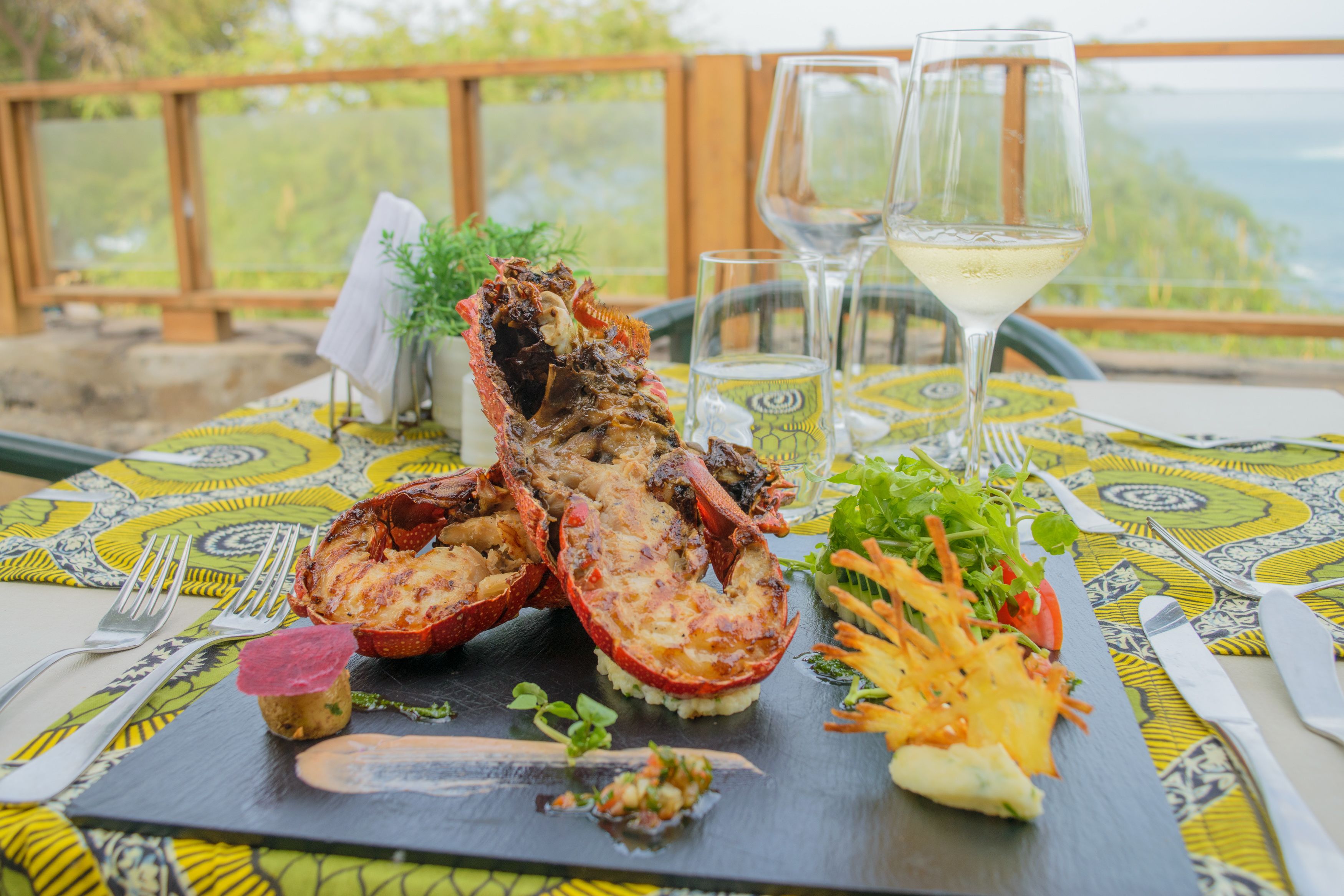 Un plato de langosta sobre una losa negra, acompañado de una ensalada y una copa de vino blanco, colocado sobre una mesa con mantel estampado.