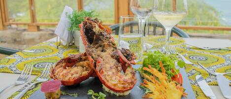 Un plato de langosta sobre una losa negra, acompañado de una ensalada y una copa de vino blanco, colocado sobre una mesa con mantel estampado.