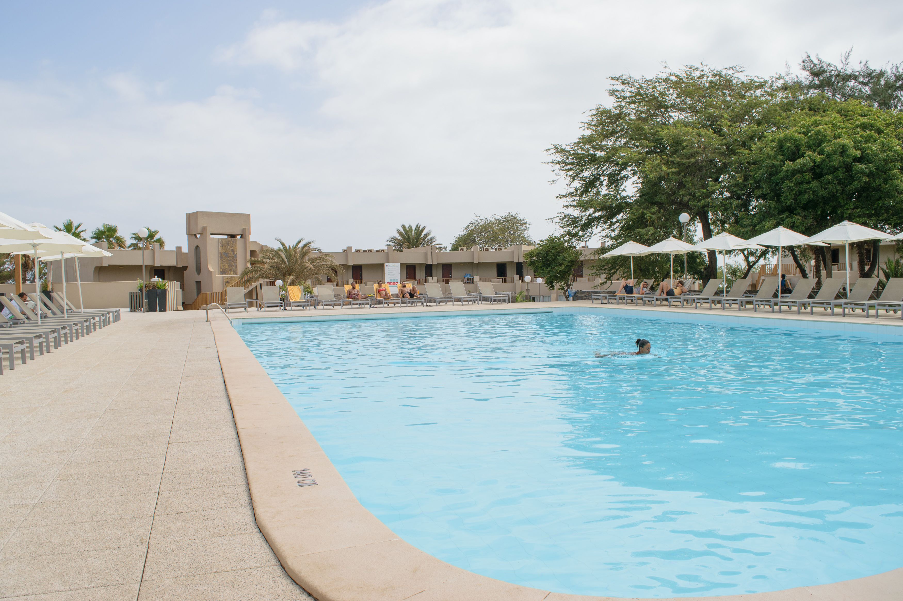 Une piscine entourée de palmiers et d’un bâtiment à l’architecture moderne, avec des chaises longues et des parasols à proximité.