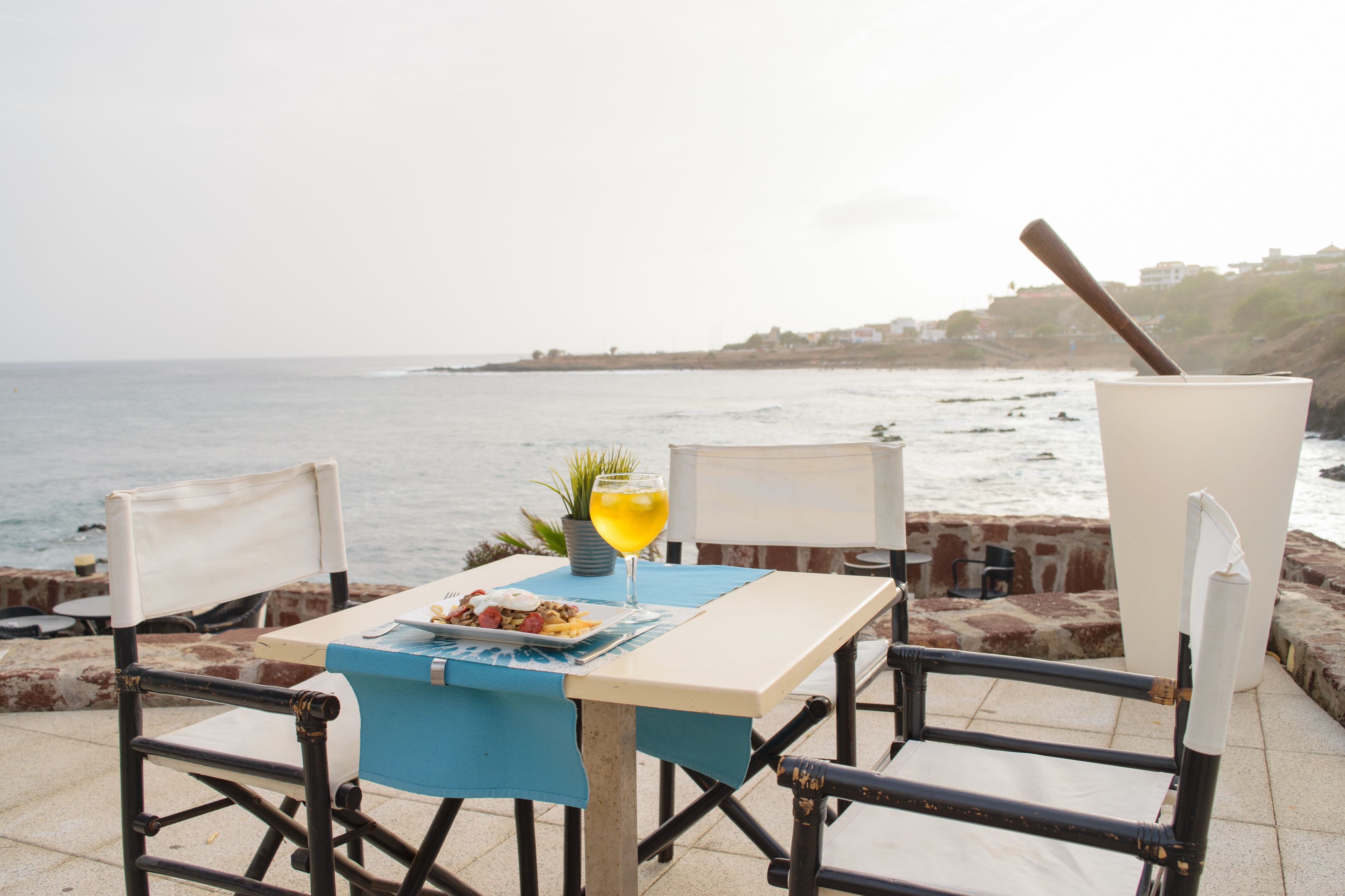 Una mesa con mantel azul, un vaso de bebida amarilla y un plato de comida, con el mar y una costa rocosa de fondo.