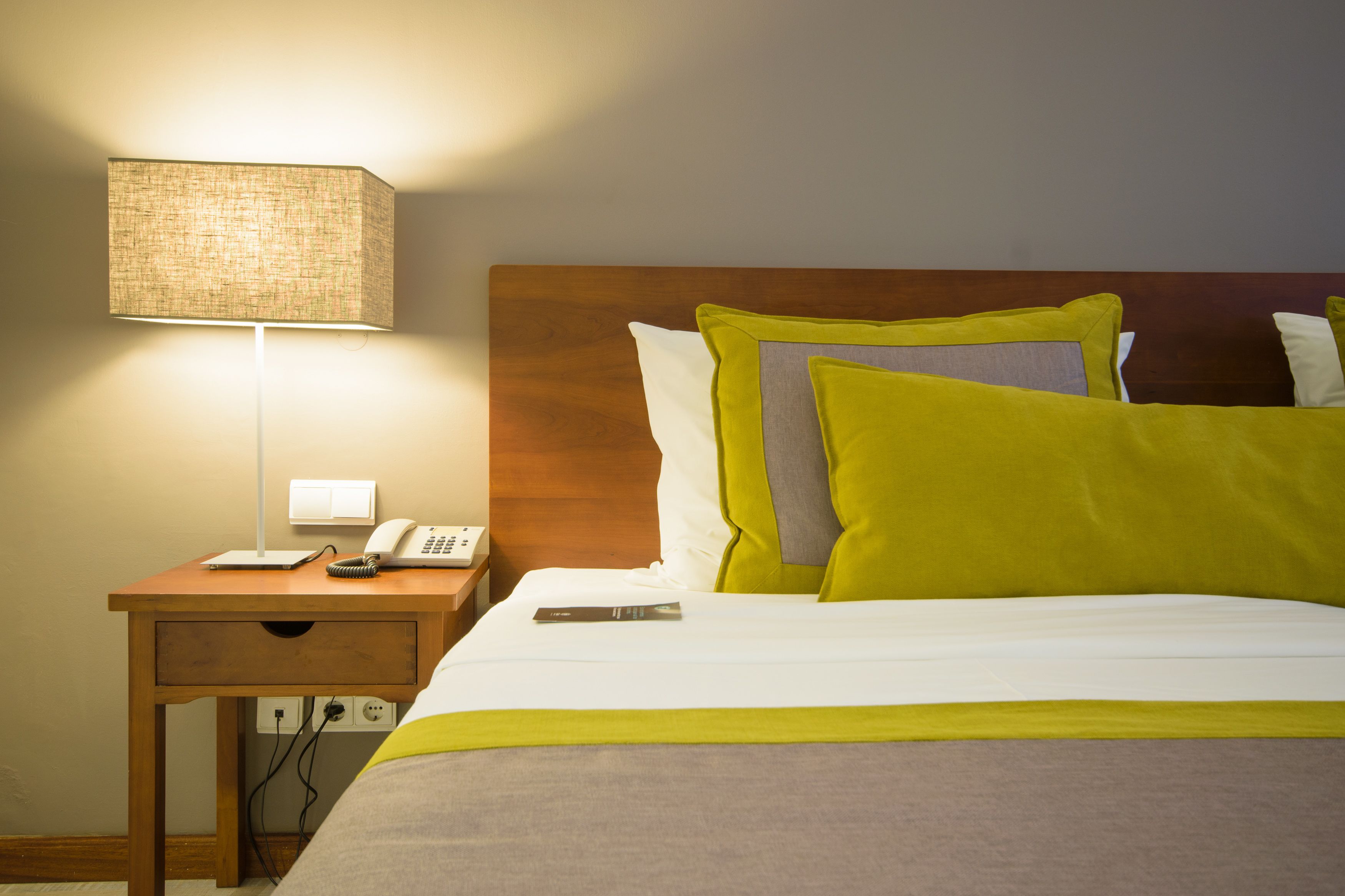 Une chambre d’hôtel avec une tête de lit en bois, une table de chevet avec une lampe, un téléphone et un lit recouvert d’une courtepointe blanche et grise, agrémenté de plusieurs oreillers.