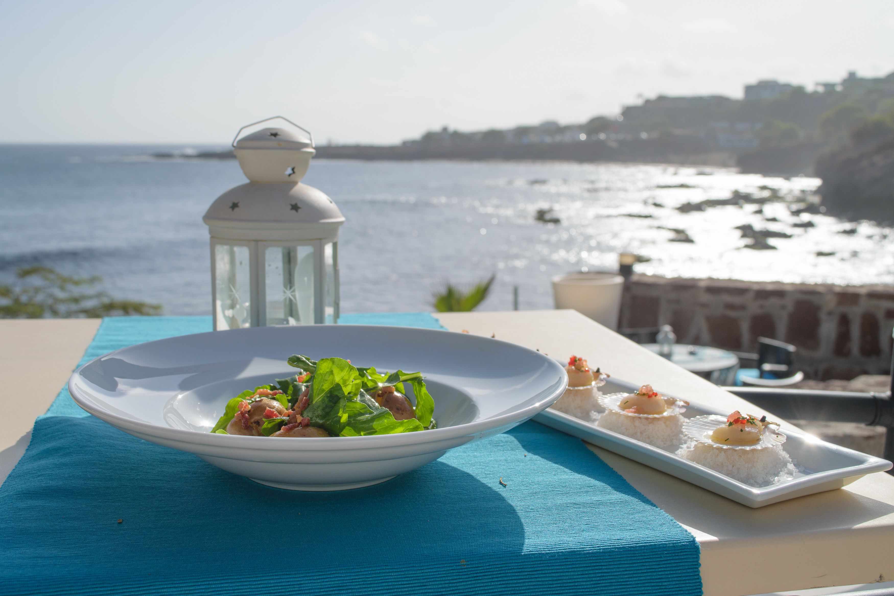 A table with a bowl of salad and a plate of appetizers, overlooking a coastal view with a lantern and a stone wall.