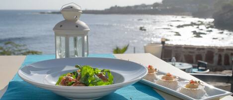 Een tafel met een saladeschaal en een bord met voorgerechten, met uitzicht op de kust, een lantaarn en een stenen muur.