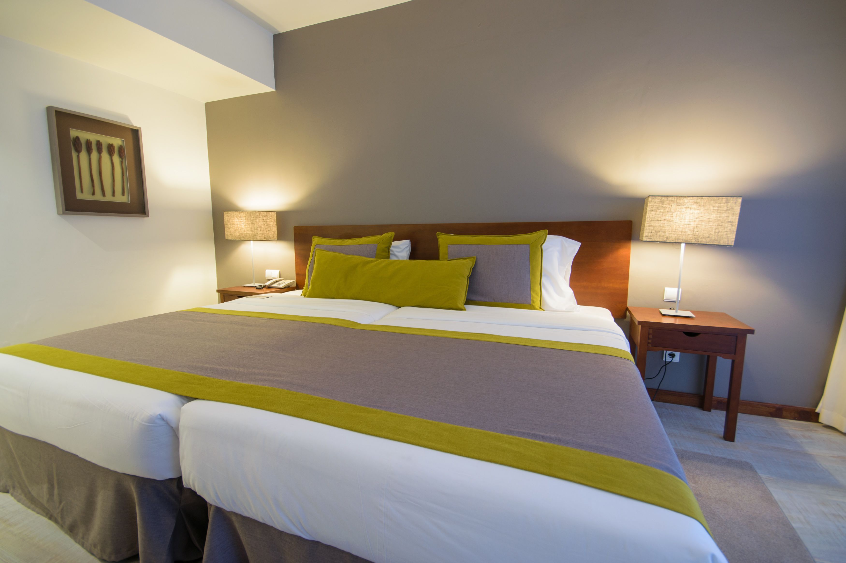 A neatly made bed with a grey and yellow bedspread, two bedside lamps, and a framed picture on the wall.