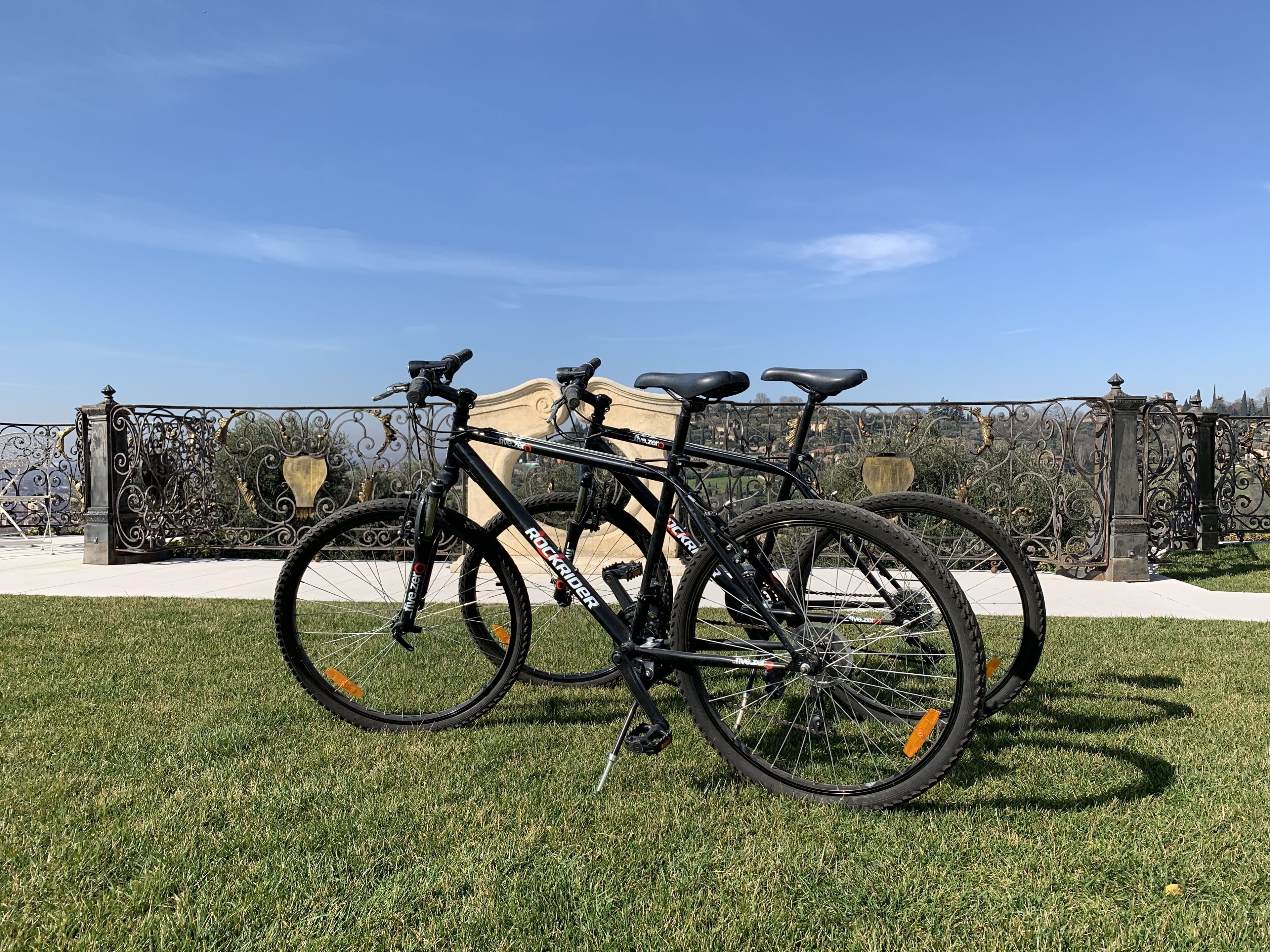 Two bicycles parked on a grassy area with a decorative fence and a clear blue sky in the background.