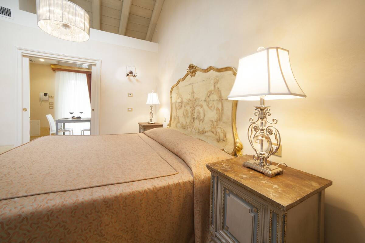 A hotel room with a large bed, a nightstand with a lamp, a headboard with a decorative design, and a view of the bathroom through an open door.