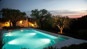 Ein rechteckiger Pool mit einer gefliesten Kante, umgeben von einer niedrigen Steinmauer. Bäume mit beleuchteten Stämmen und eine Bank liegen in der Nähe des Pools.