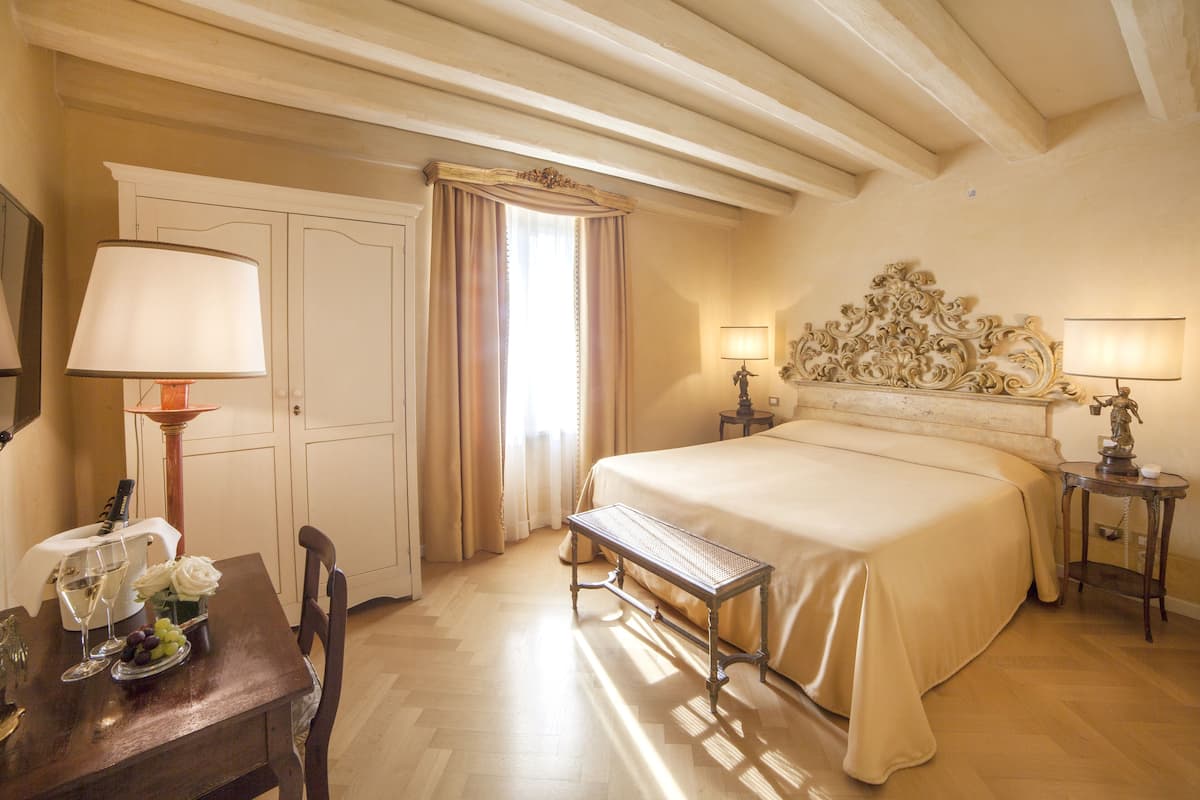 A hotel room with a bed, bedside tables, a bench, a desk with a lamp, and a window with curtains.
