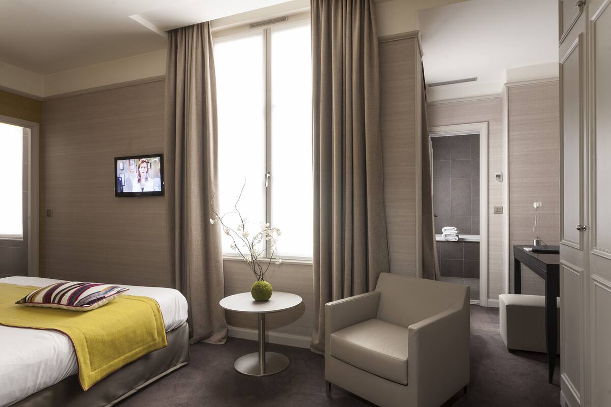 A hotel room with a bed, a chair, a small table with a vase, a TV, and a bathroom visible through an open door.