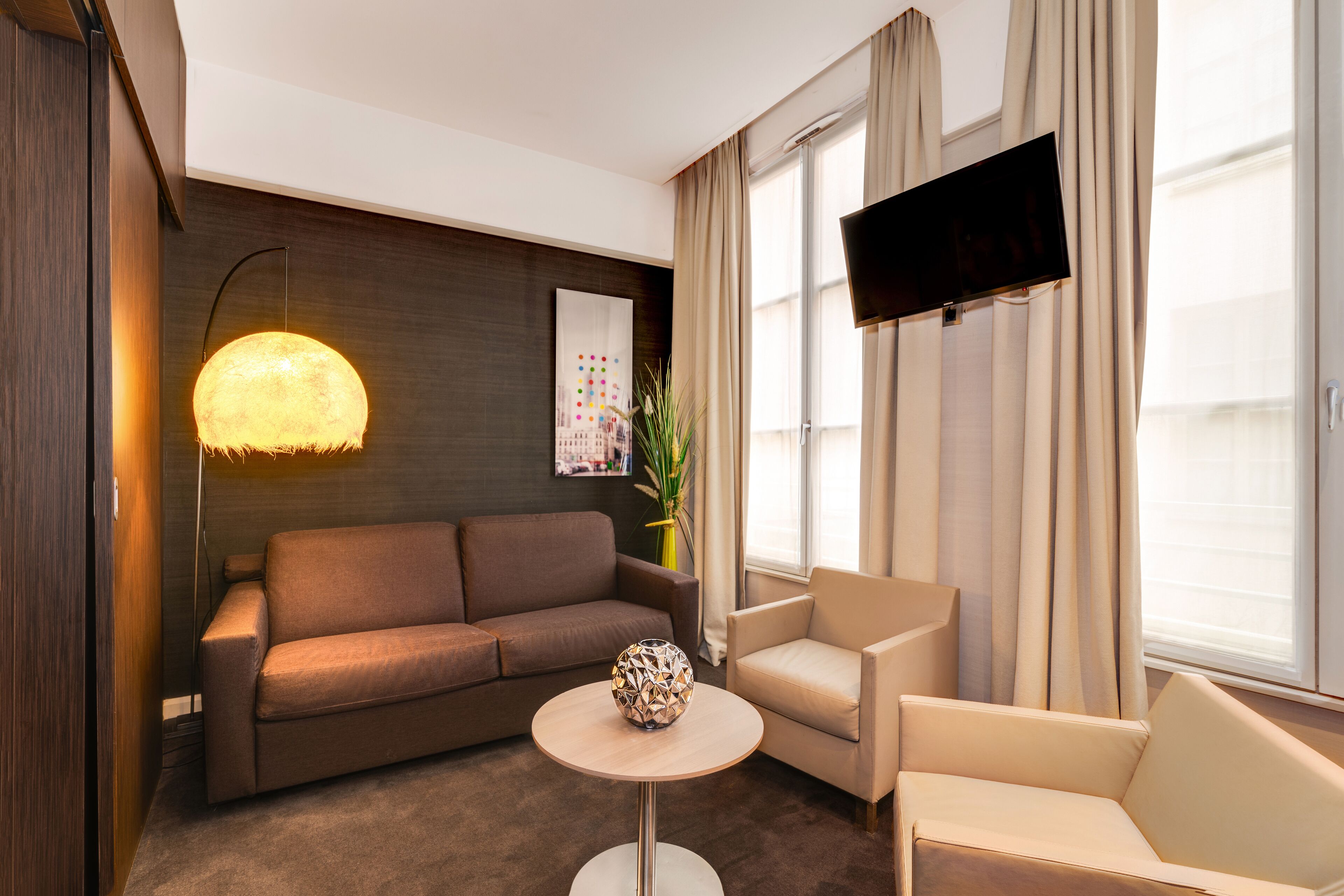 A modern living room with a brown sofa, a beige armchair, a round table with a decorative vase, a wall-mounted TV, and a large window with curtains.