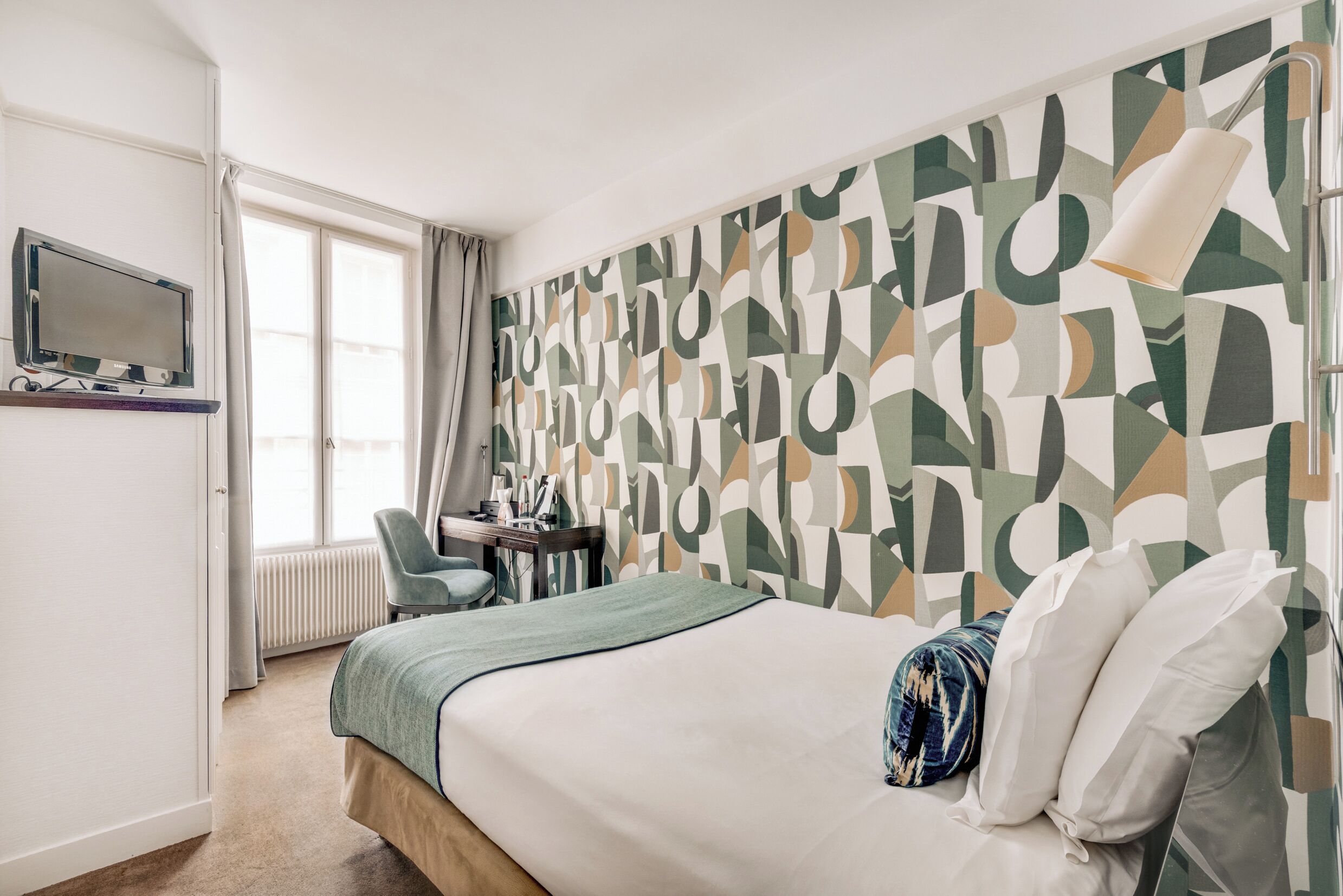 Habitació d'hotel amb un llit, una taula de treball amb una cadira, una televisió i una paret amb un disseny decoratiu.