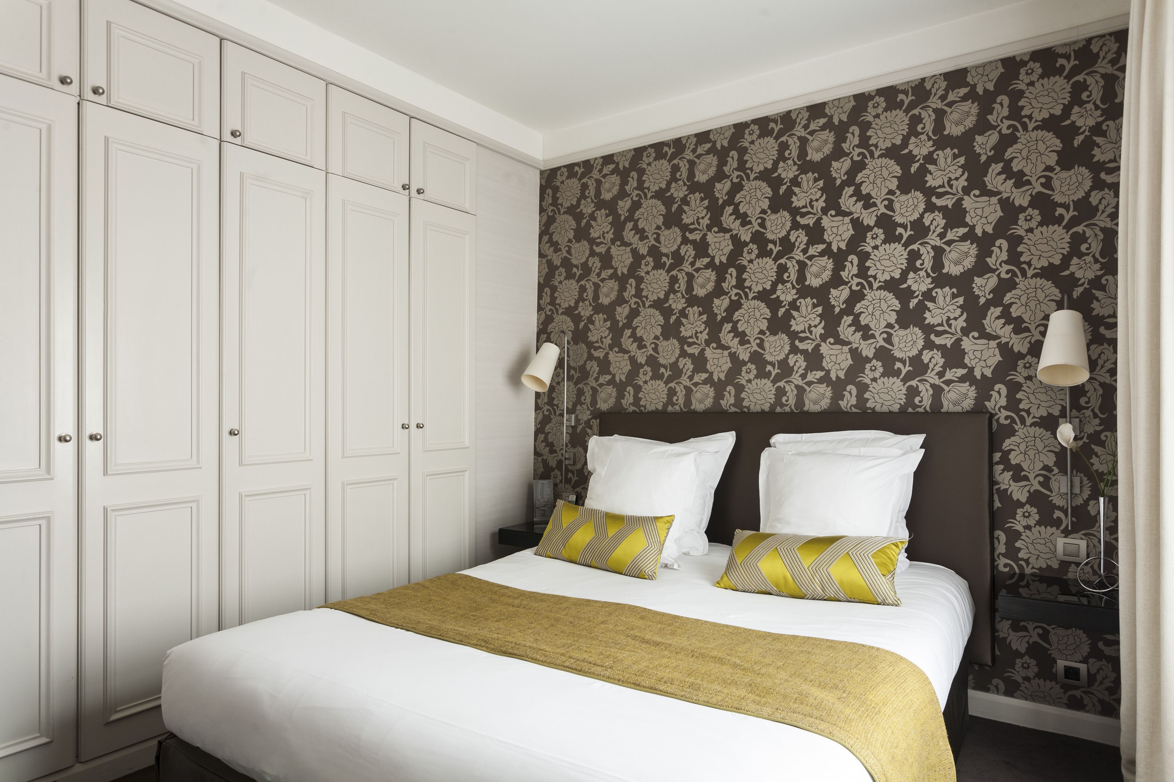 A neatly made bed with white bedding and two decorative pillows, a bedside table with a lamp, and a wardrobe with multiple doors.