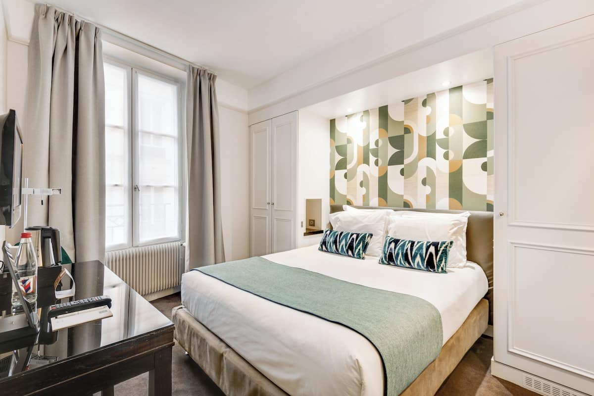 A hotel room with a bed, a desk, a chair, a window with curtains, a radiator, and a wall with a geometric pattern.