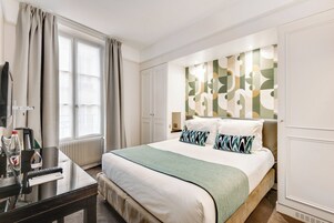 Une chambre d’hôtel comprenant un lit, un bureau, une chaise, une fenêtre avec des rideaux, un radiateur et un mur orné d’un motif géométrique.